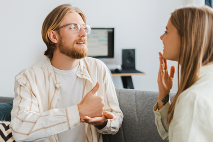 Zwei Personen unterhalten sich in Gebärdensprache. Beide sind blond, die Person links trägt eine Brille. Sie sitzen auf einem grauen Sofa in einem Wohnzimmer.