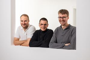 Three men leaning against a white wall, smiling