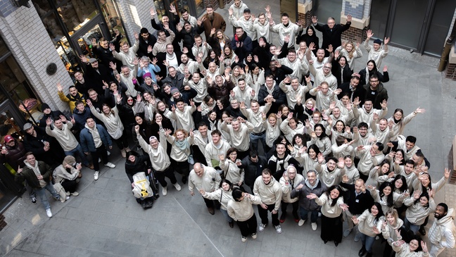 “Large group of smiling Eye-Able team members standing in a courtyard, waving up at the camera, many wearing matching light-colored hoodies