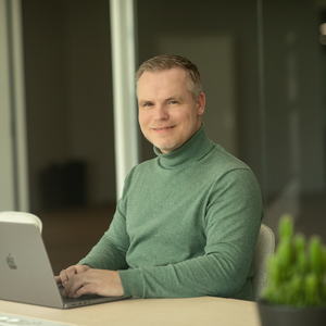 A smiling man in a green sweater sitting on a laptop.