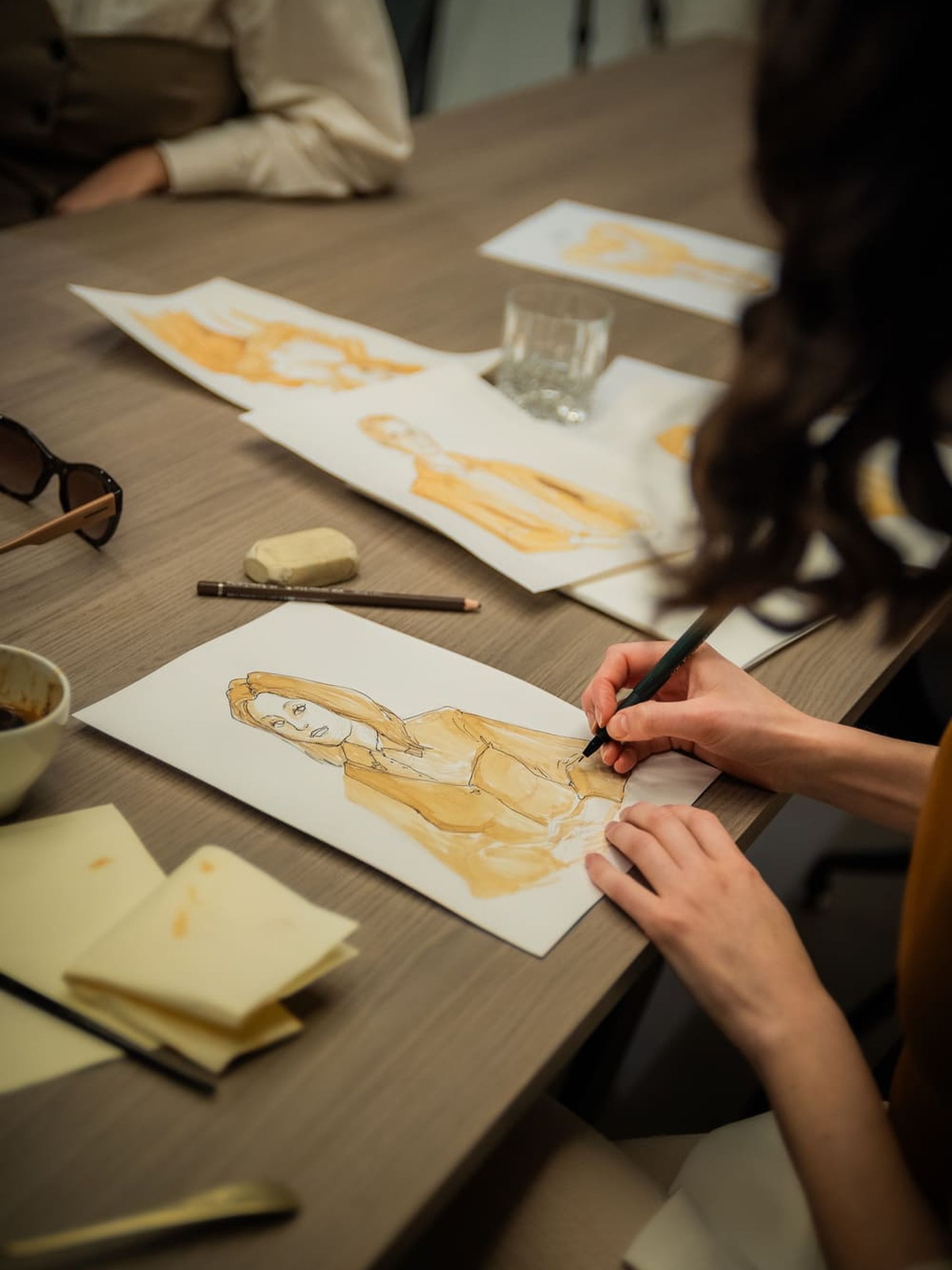 A person sketches a portrait using a brush at a wooden desk, surrounded by papers, a glass, and art supplies.