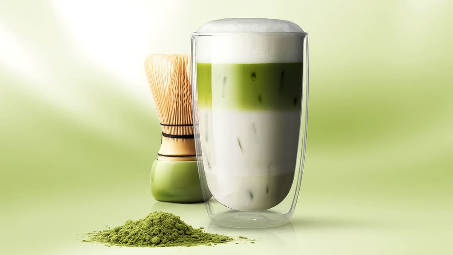 A glass of layered green matcha latte with white foam, placed next to a bamboo whisk and a pile of matcha powder on a soft green background.