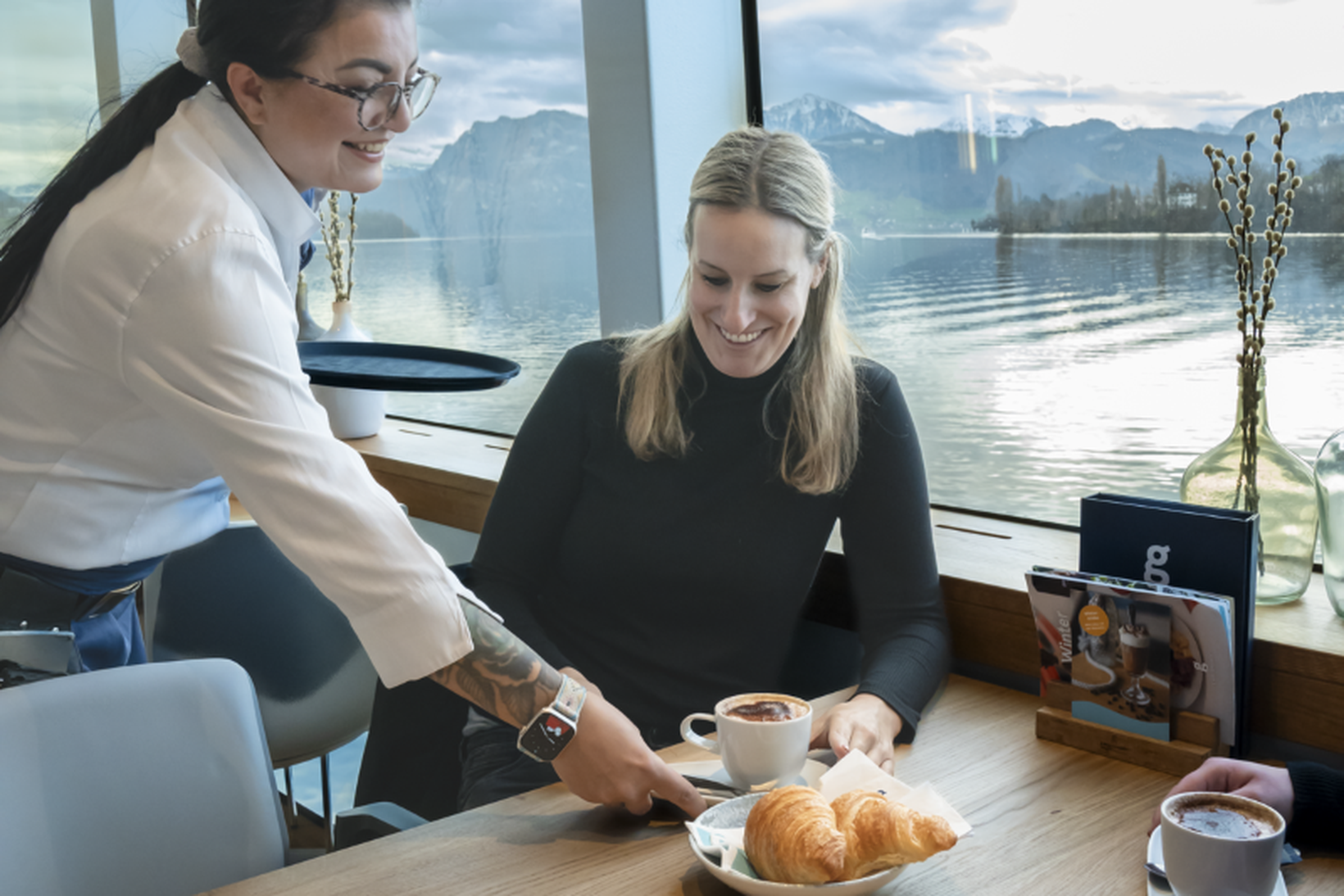 A woman in a cafe receives coffee and croissants from a server. The setting overlooks a lake with mountains in the background.