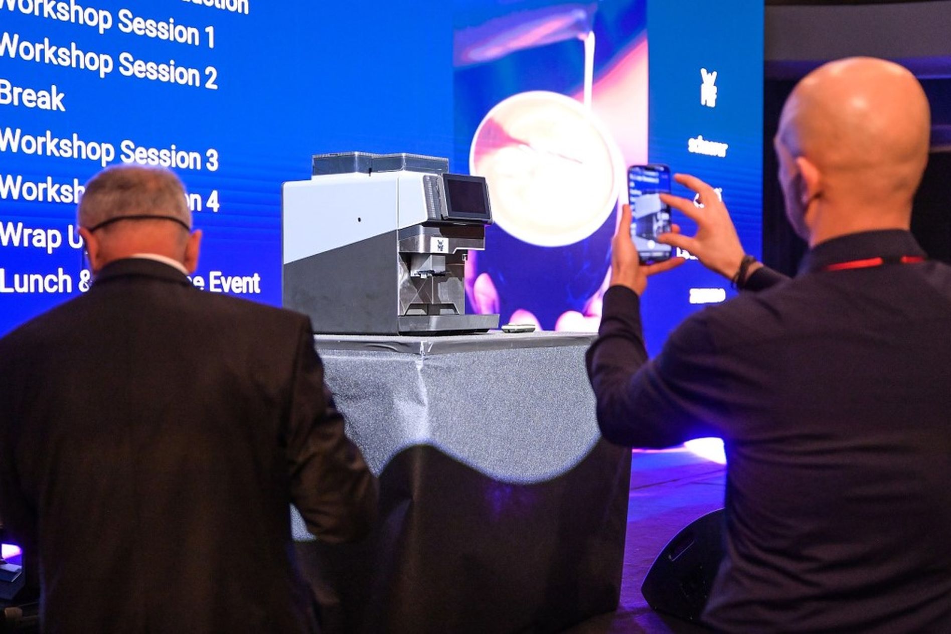 Two people photographing a professional coffee machine on display at a conference, with a large schedule projected in the background.