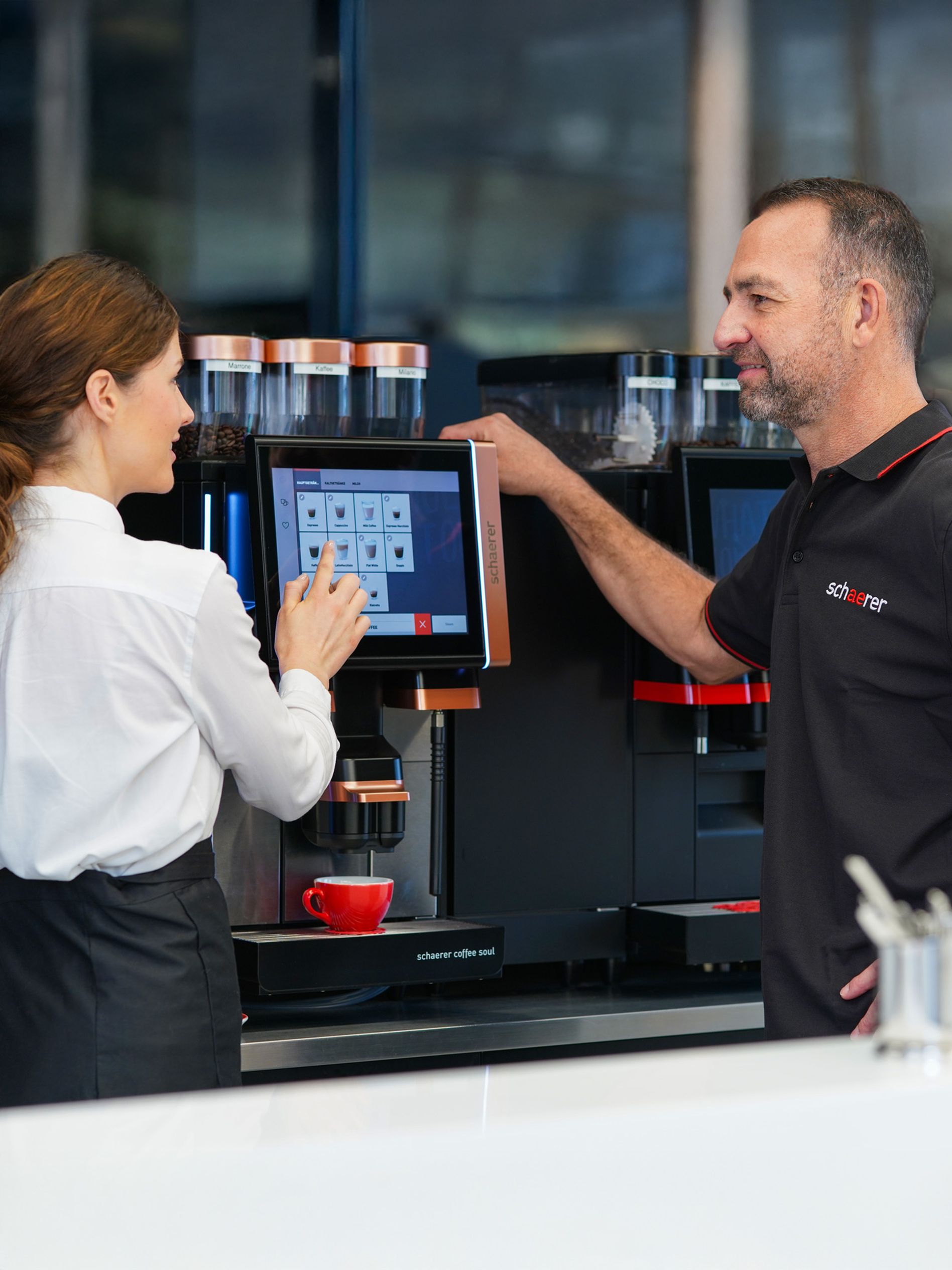 A woman and a man operate a touchscreen coffee machine in a modern setting. A red cup is placed on the machine's tray.