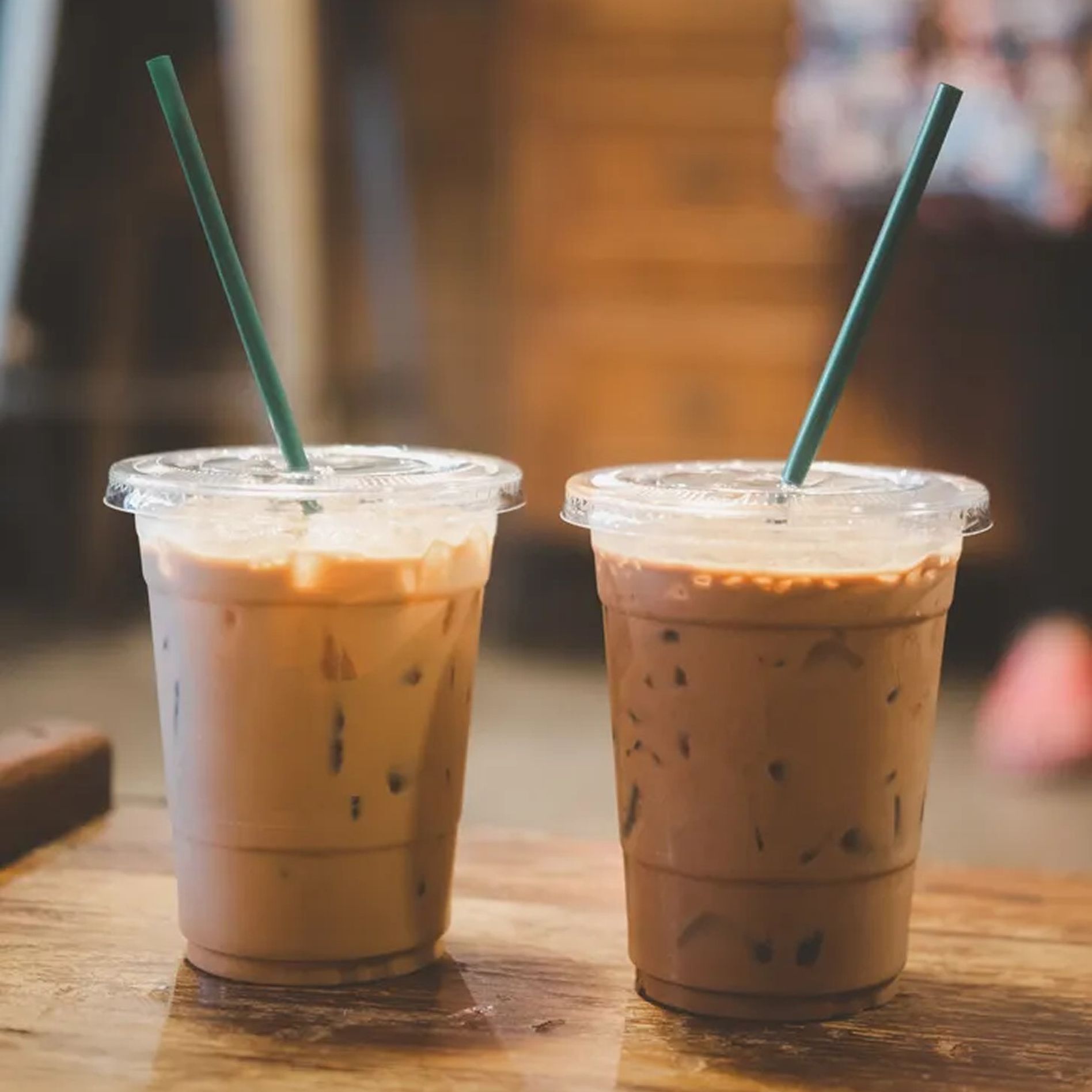 Two iced coffee drinks with green straws sit on a wooden table. The drinks are in clear plastic cups with lids, filled with ice cubes.