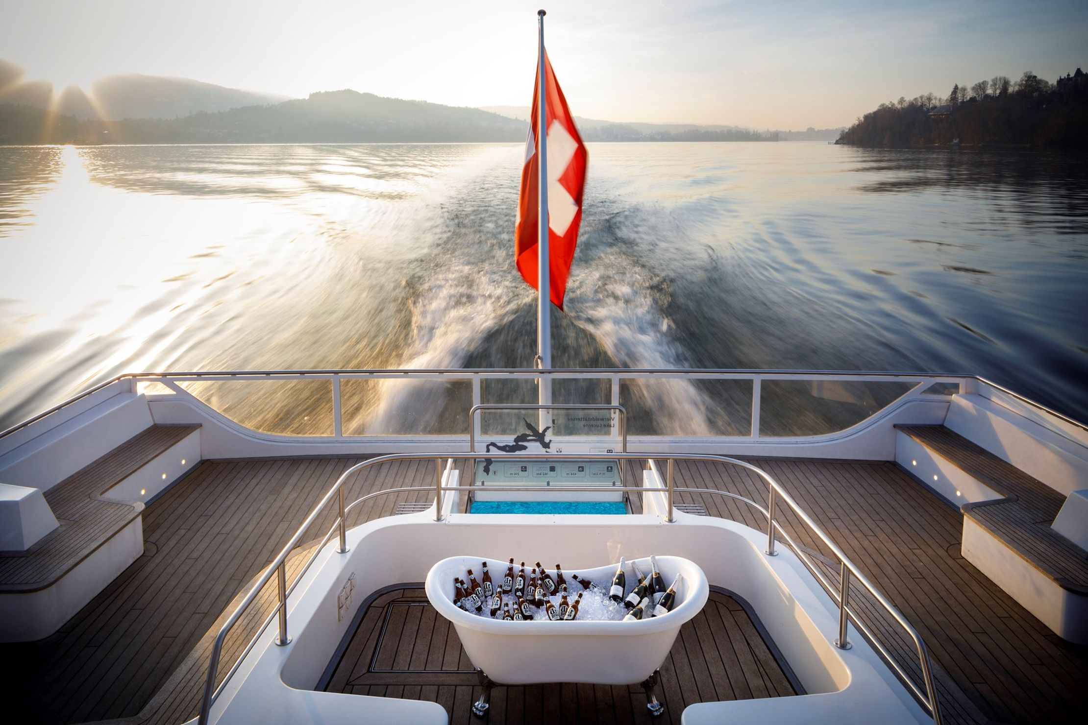 View from a boat's deck with a Swiss flag, a tub filled with ice and drinks, and a wake trailing behind on a calm lake at sunset.