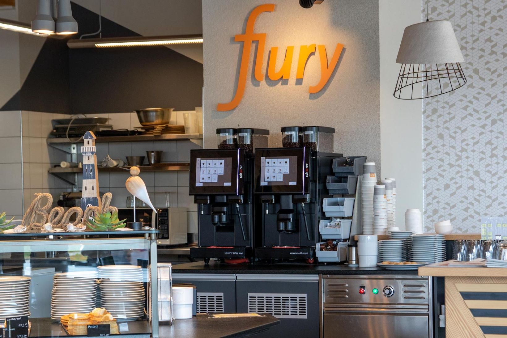 Coffee shop interior with two coffee machines, stacked cups, plates, and a decorative "Beach" sign. "Flury" logo on the wall.