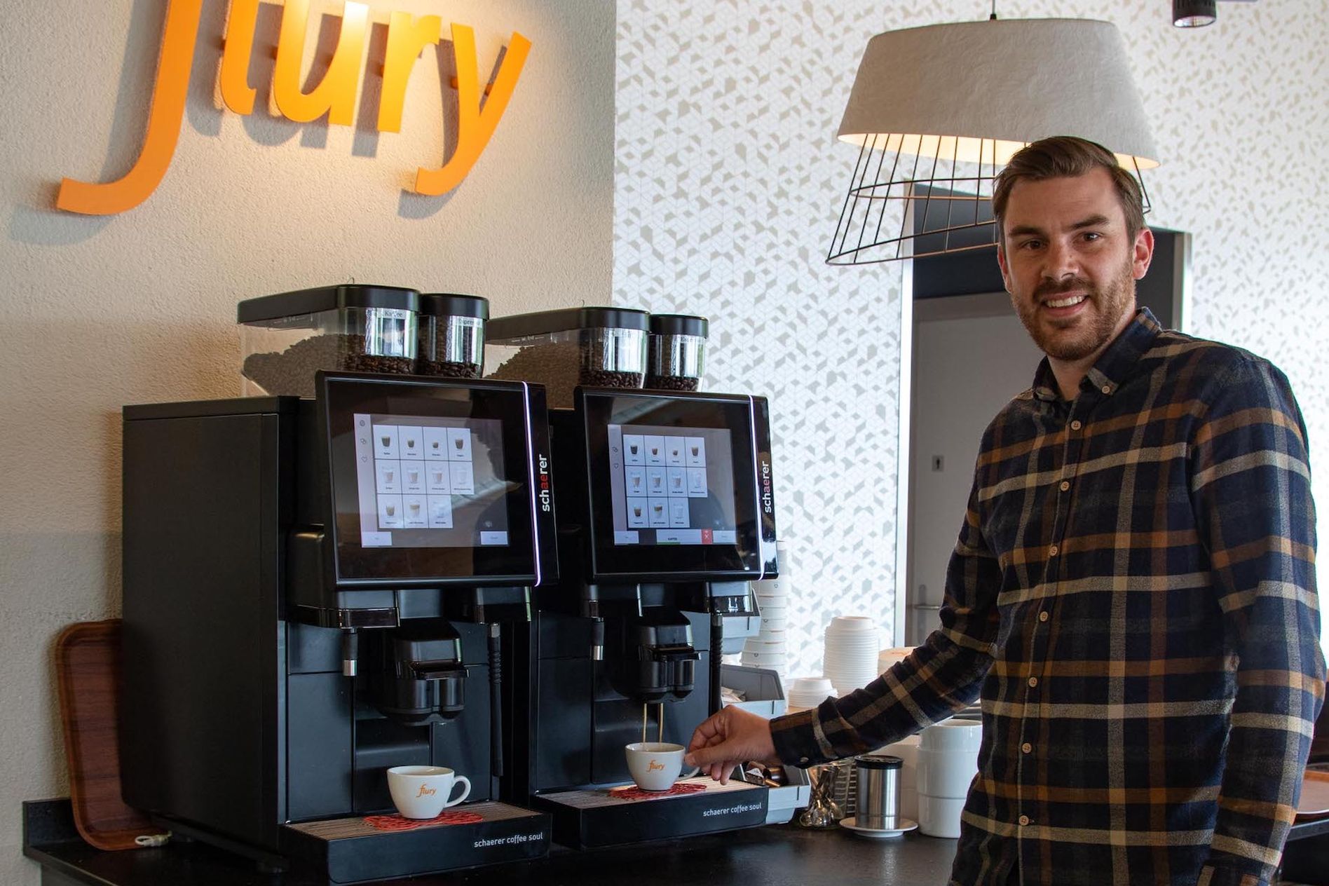 Man in a plaid shirt using a modern coffee machine in a café with "Flury" sign. Two cups are placed under the dispensers.