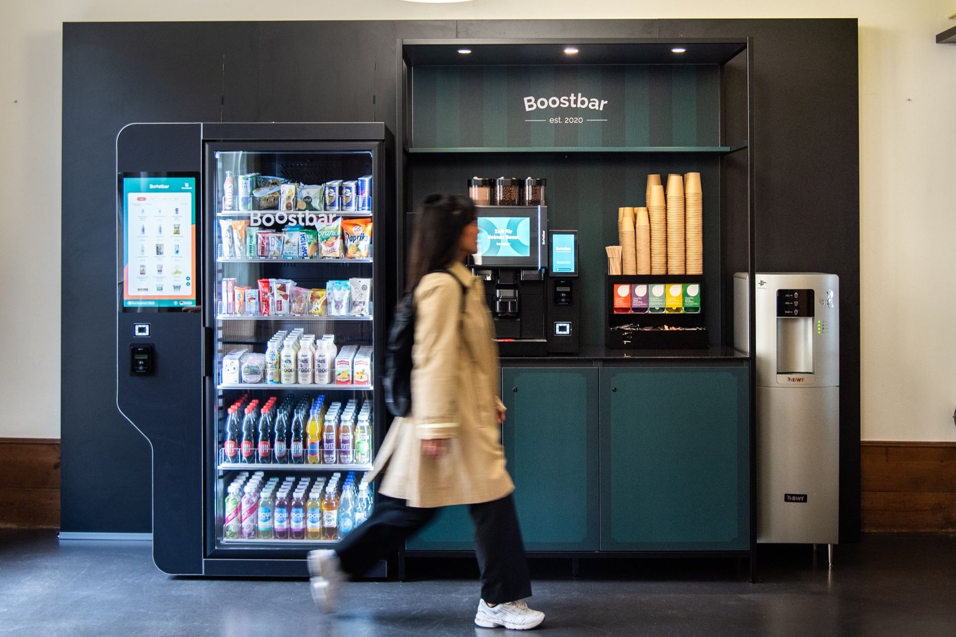 A person walks past a vending machine and coffee station labeled "Boostbar," featuring snacks, drinks, and a coffee dispenser.