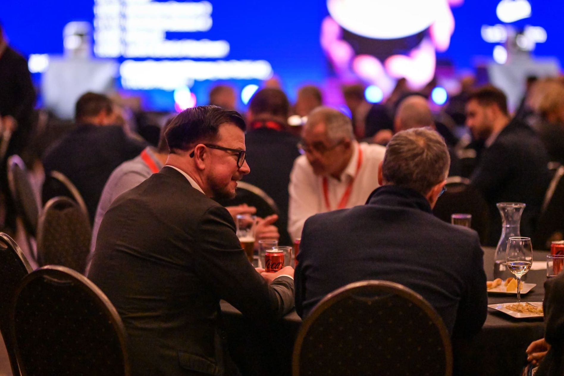 People sitting at round tables in a conference setting, engaged in conversation with a blurred blue presentation screen in the background.