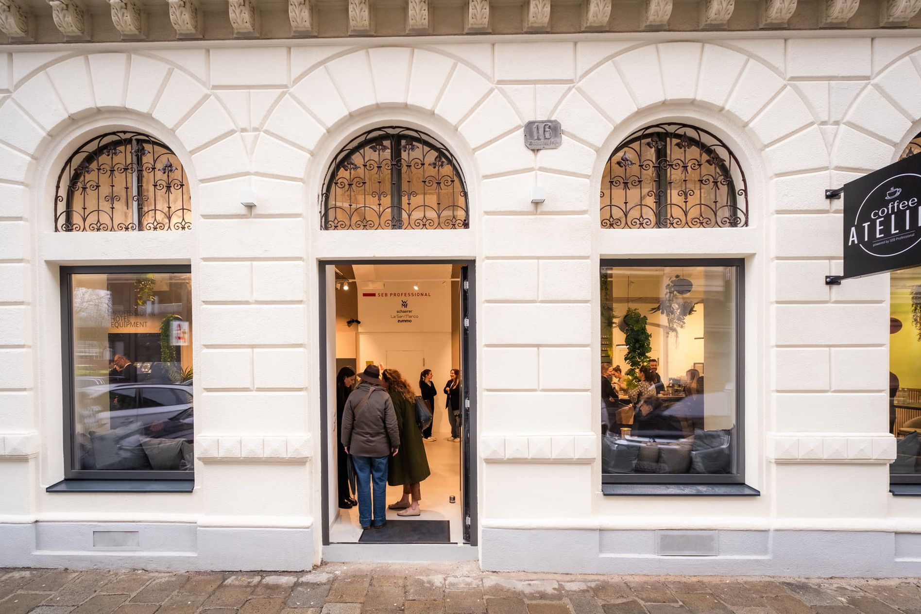 People entering a stylish café with arched windows and a sign reading "Coffee Atelier" on a city street.