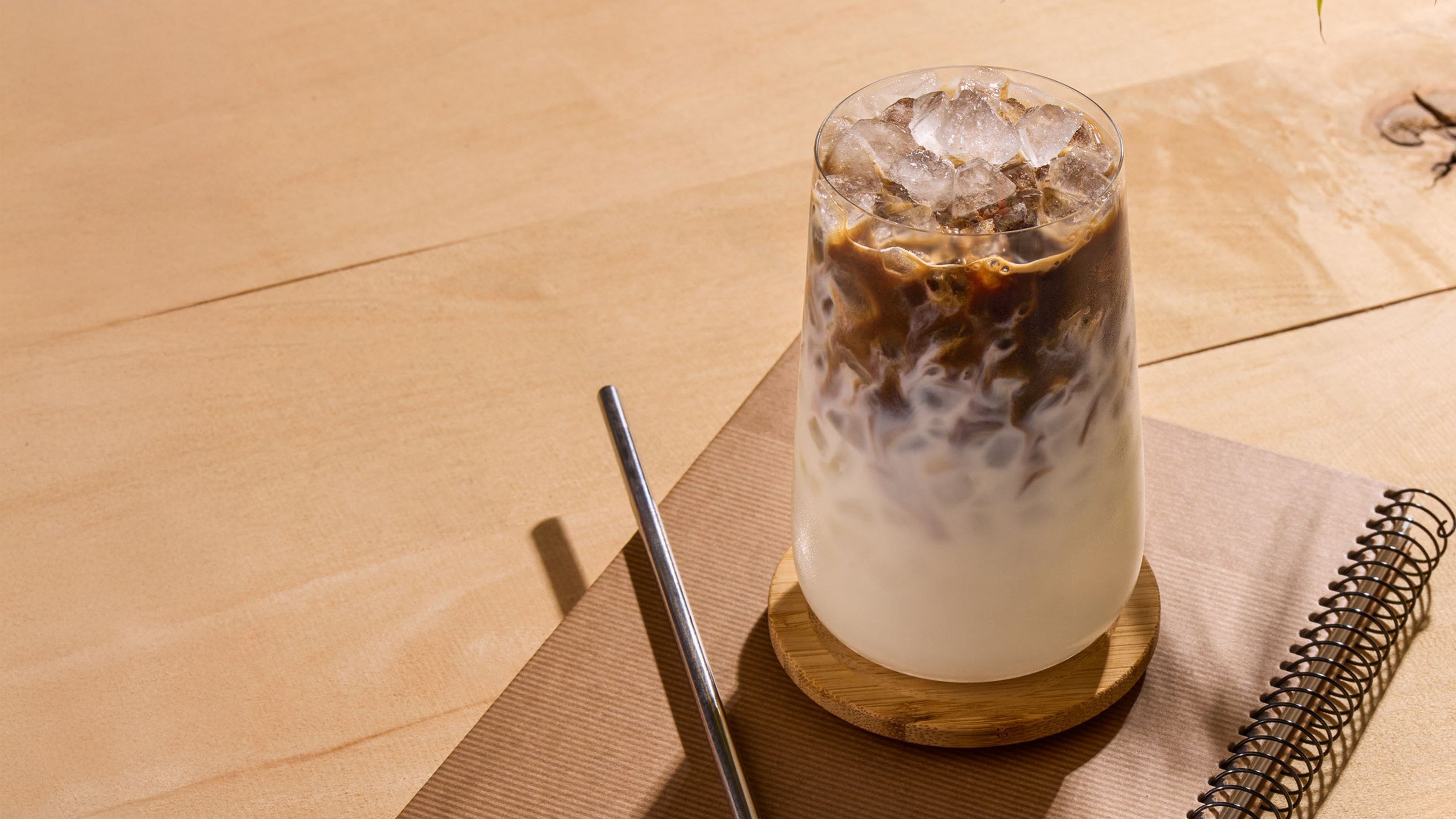 A glass of iced coffee with milk, next to a metal pen and spiral notebook on a wooden table.