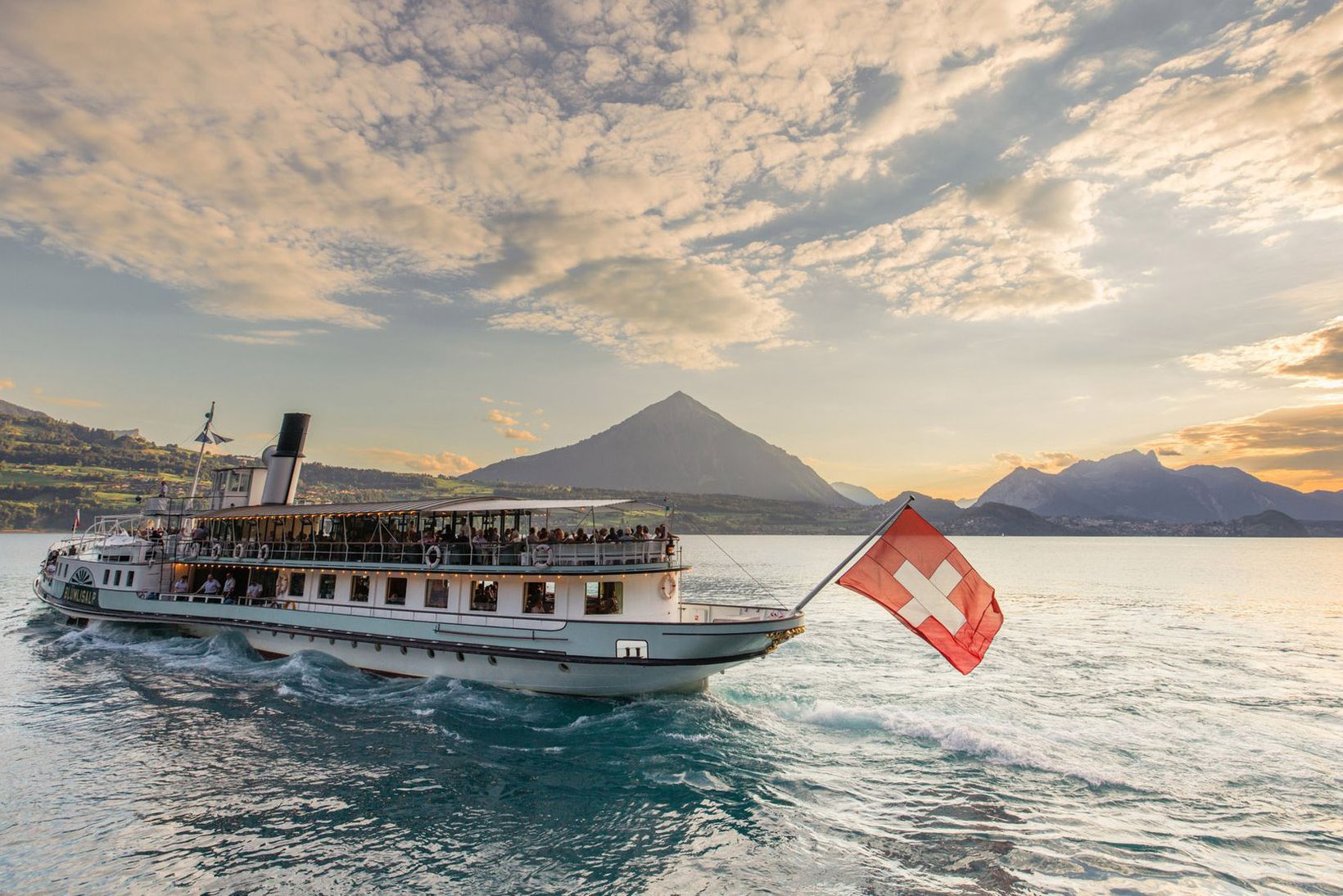 A ship with a Swiss flag sails on a lake Brienz, surrounded by mountains under a cloudy sky at sunset.