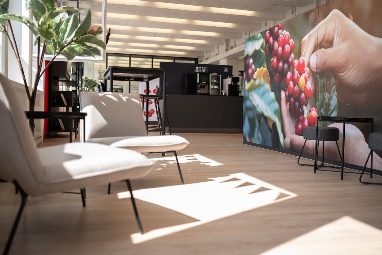 Modern office lounge with light wood flooring, gray chairs, a large mural of hands holding coffee cherries, and a potted plant by the window.