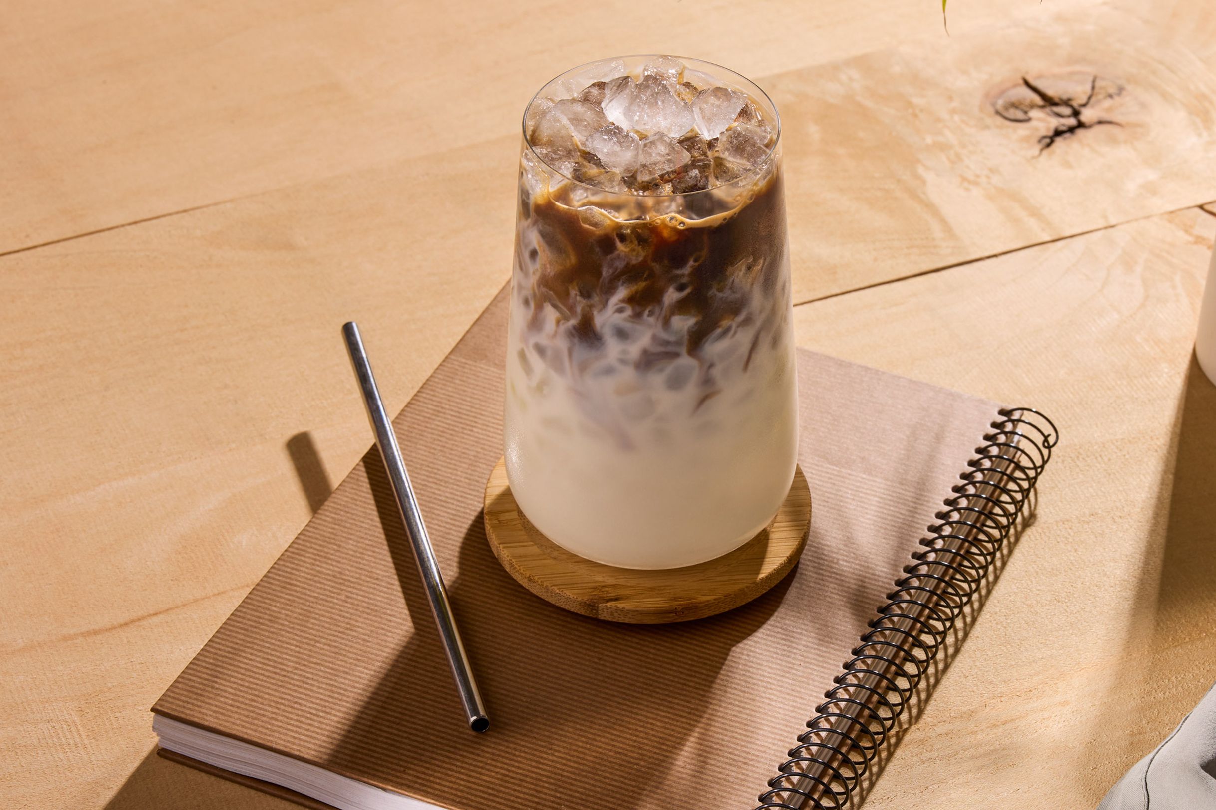 A glass of iced coffee with milk sits on a wooden coaster atop a notebook, accompanied by a metal straw, on a wooden surface.