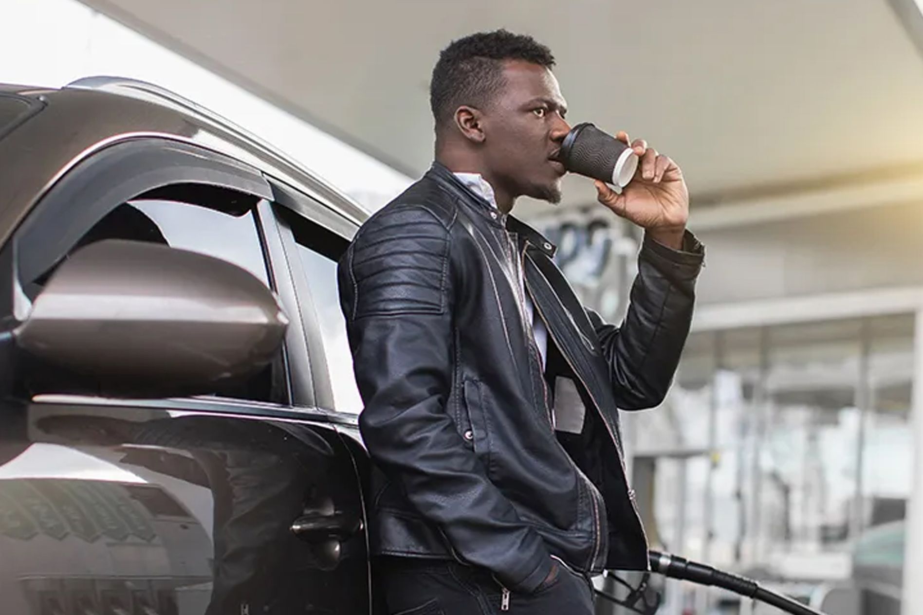 Man in a leather jacket leans against a car, drinking from a cup, at a gas station.