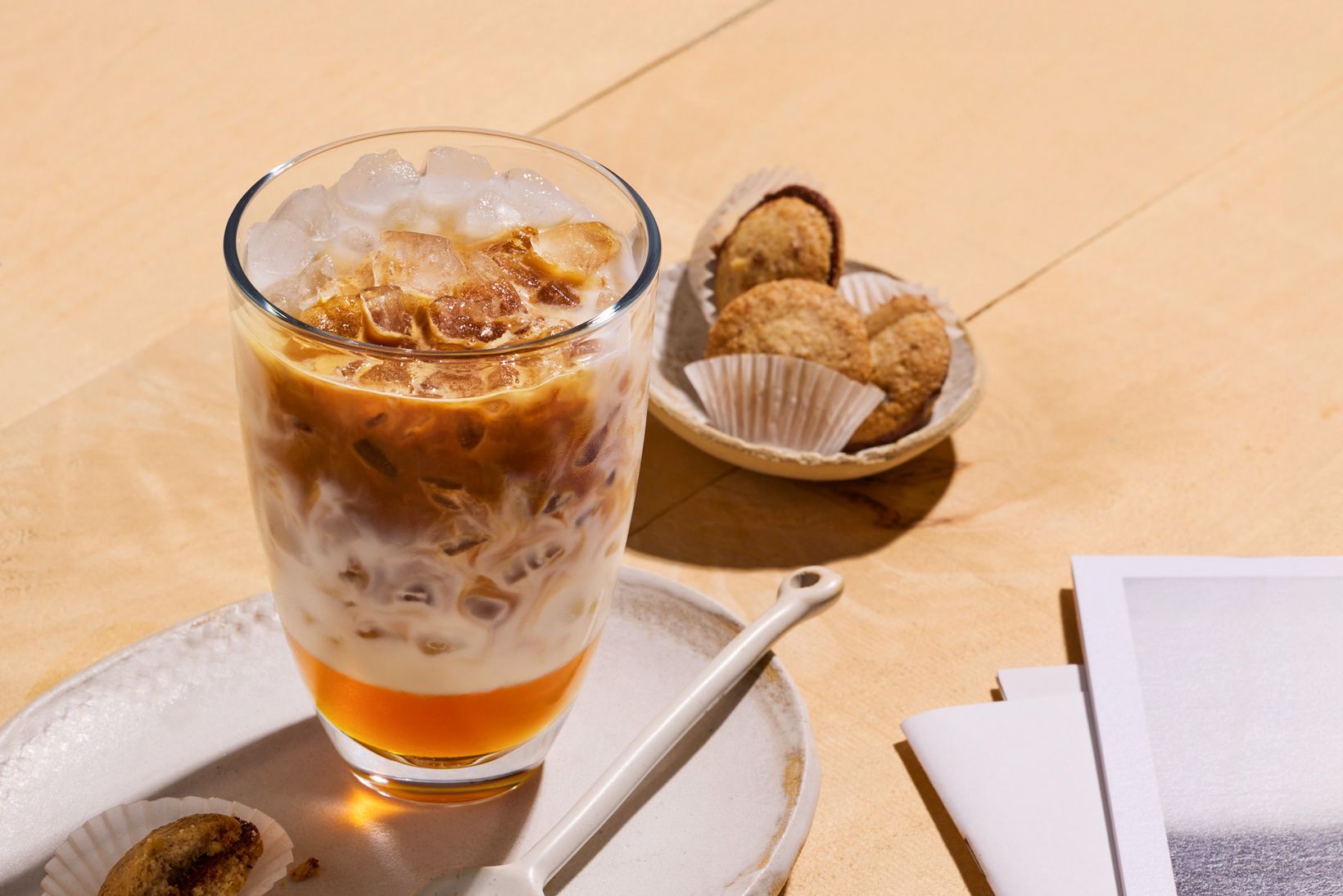 Iced coffee with milk in a glass on a plate, accompanied by cookies in paper cups and a spoon, set on a wooden surface.