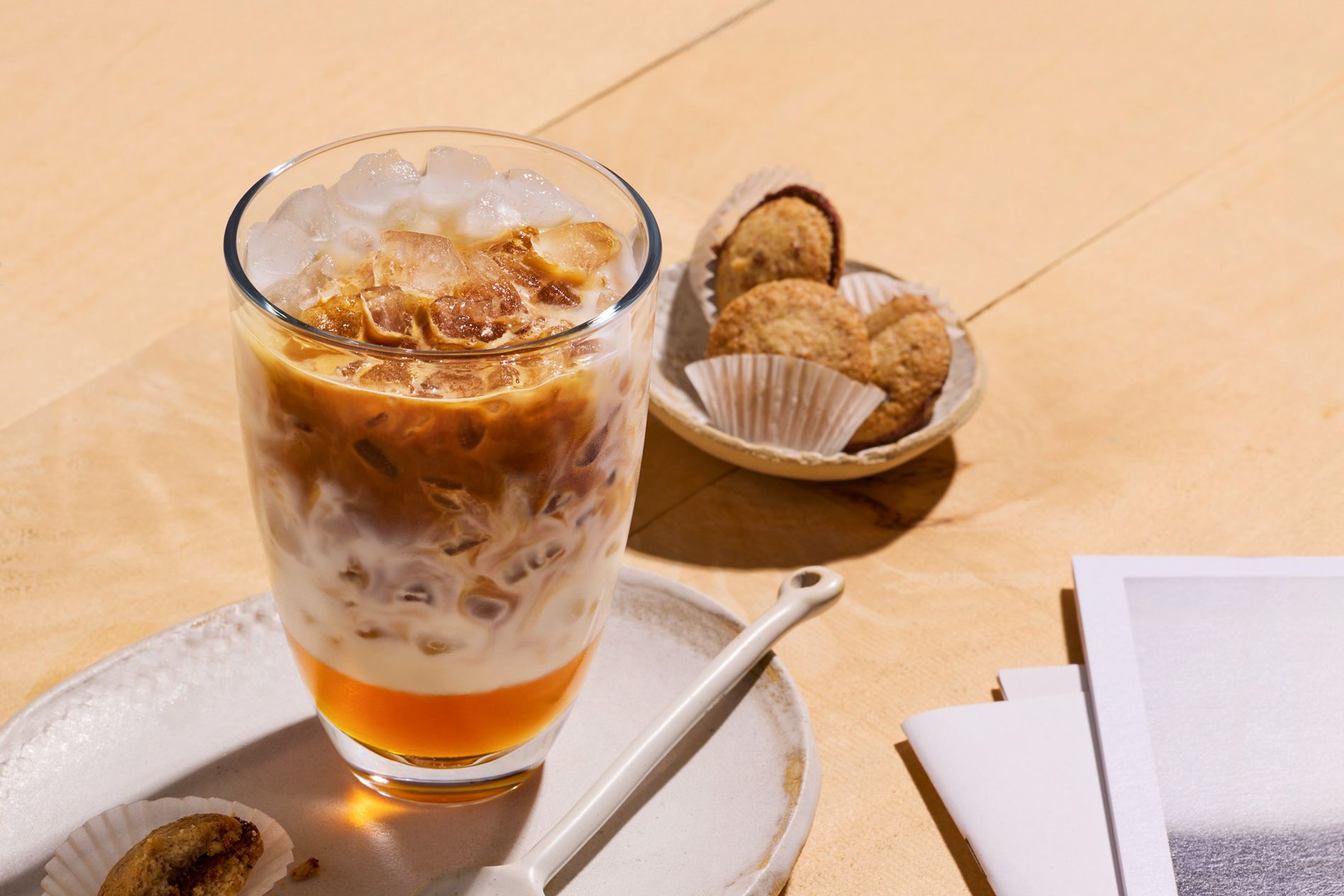 Iced coffee with milk in a glass on a plate, accompanied by cookies in paper cups and a spoon, set on a wooden surface.