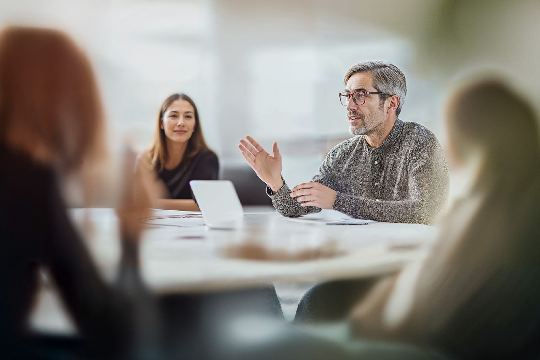 Man speaking in a meeting, gesturing with his hand, while a woman listens attentively. Blurred foreground and background elements.