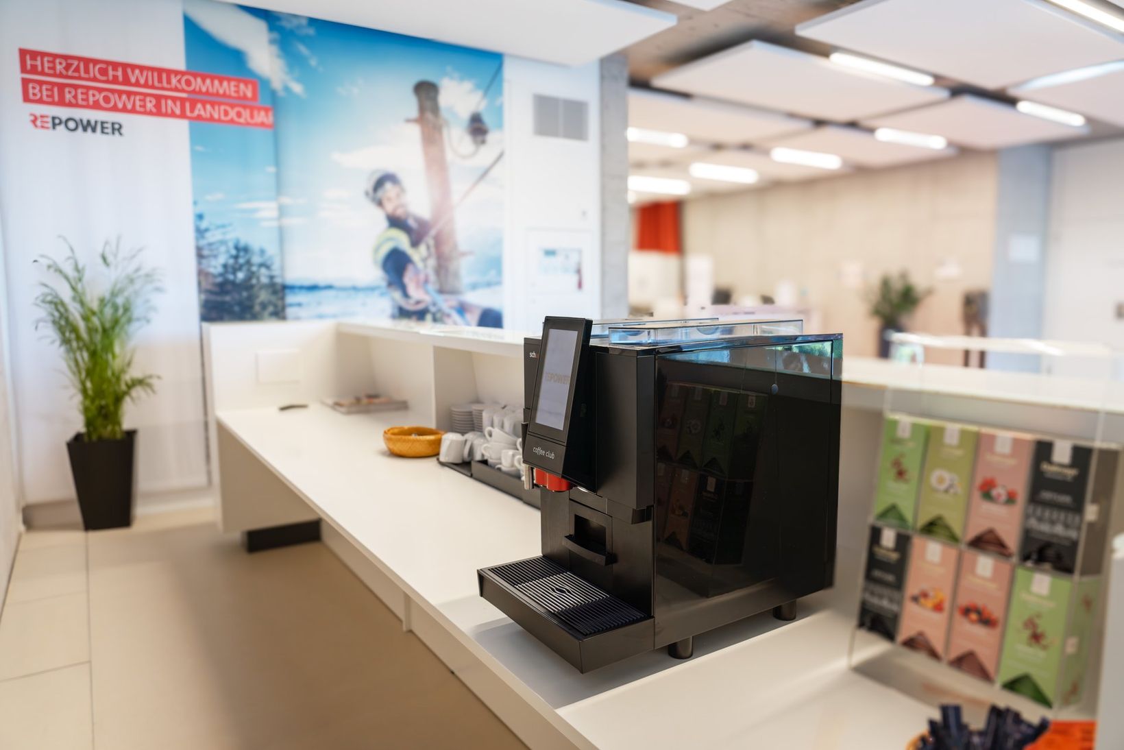 Modern office break area with a coffee machine on a counter, snack bowls, and a wall poster featuring a welcoming message.