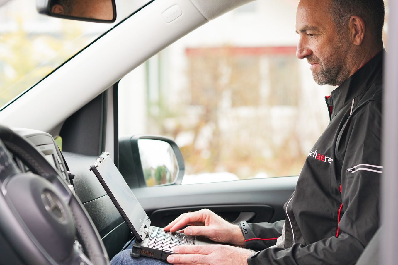 Man sitting in a car, using a laptop on his lap, wearing a black jacket with red accents, focused on the screen.