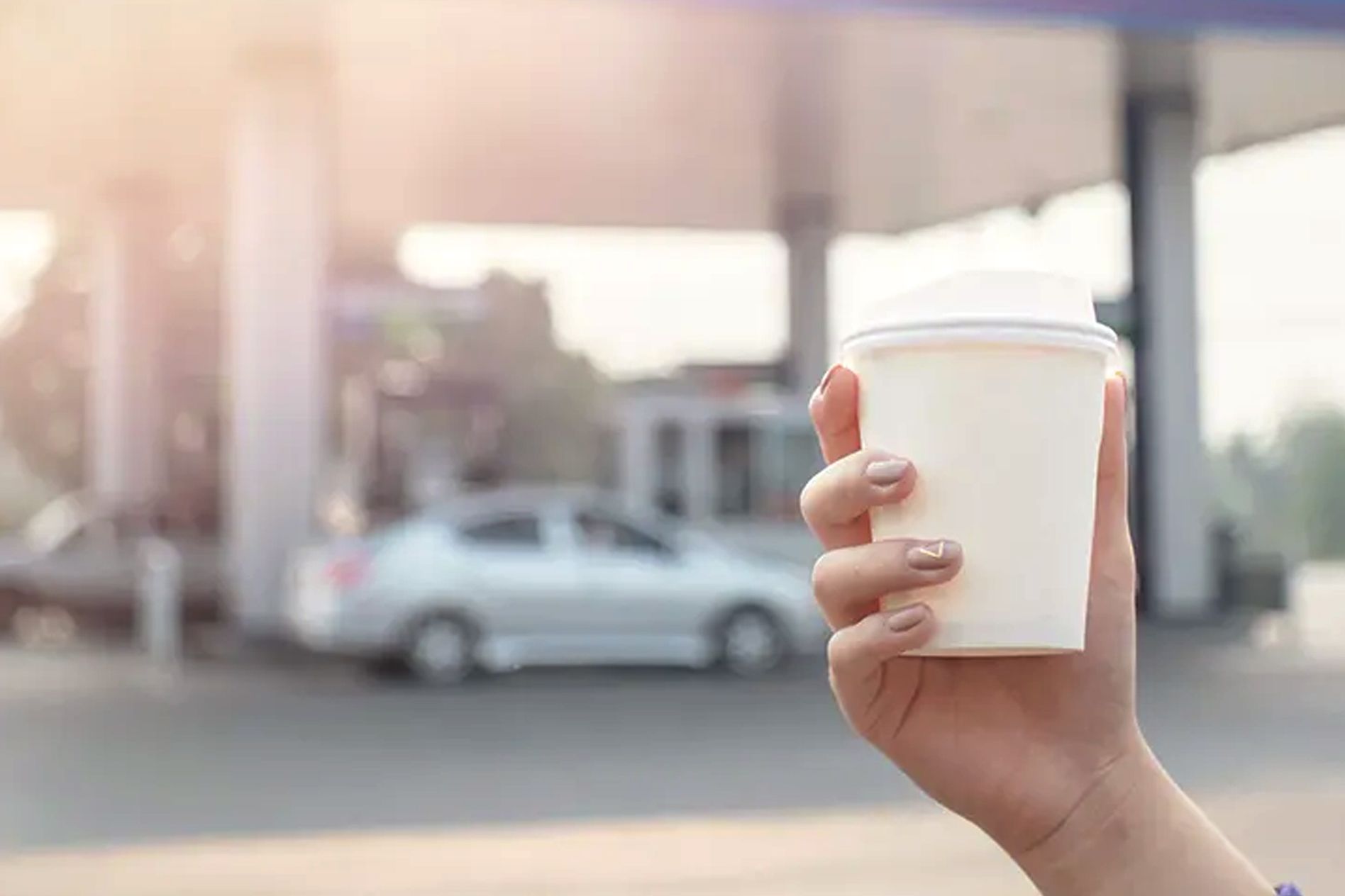 Hand holding a white paper coffee cup in front of a gas station with a blurred silver car in the background.