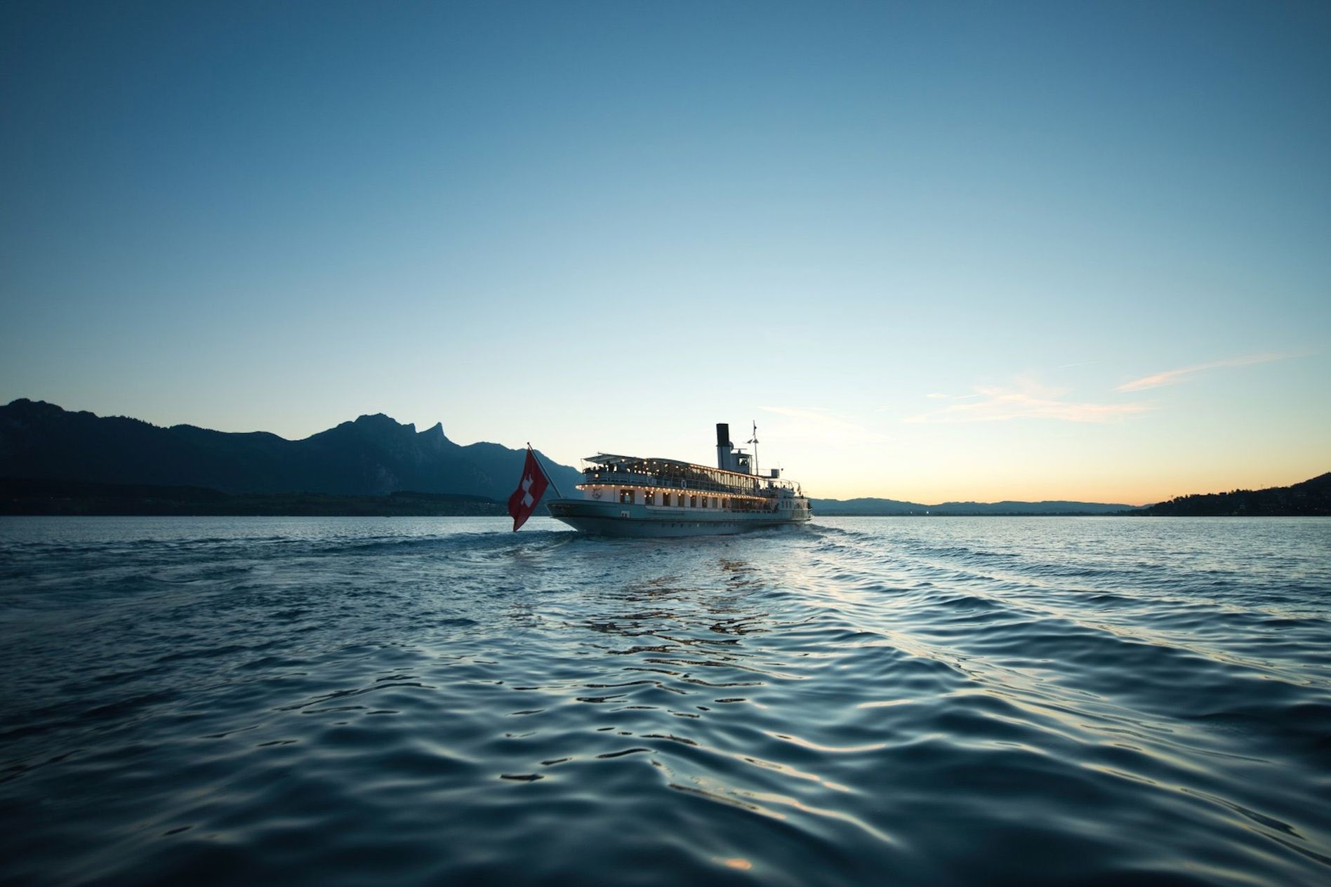 A boat with a Swiss flag sails on Lake Brienz at sunset, with silhouetted mountains in the background