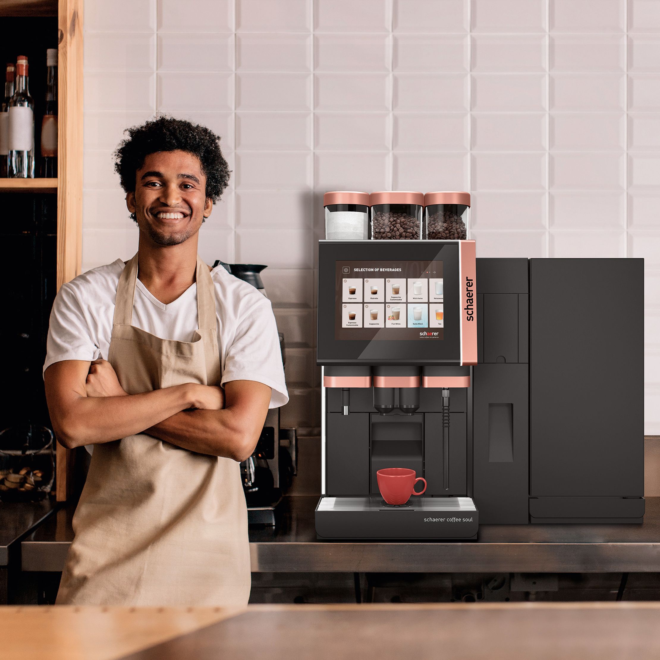 Barista in apron smiles beside a modern coffee machine with a red cup, set against a tiled wall in a cozy cafe.