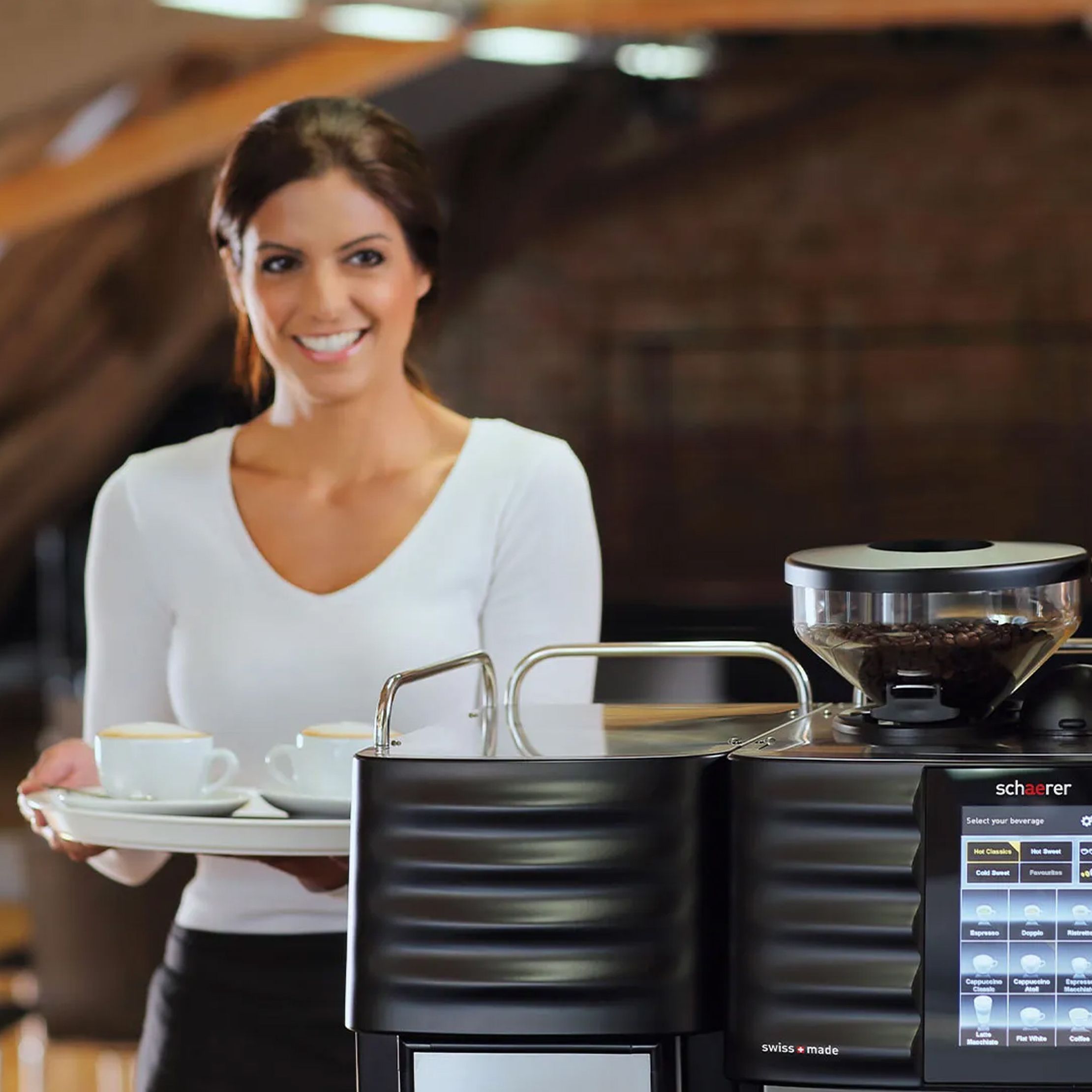 Woman in a white shirt holding a tray with cups stands beside a modern coffee machine in a cozy setting.