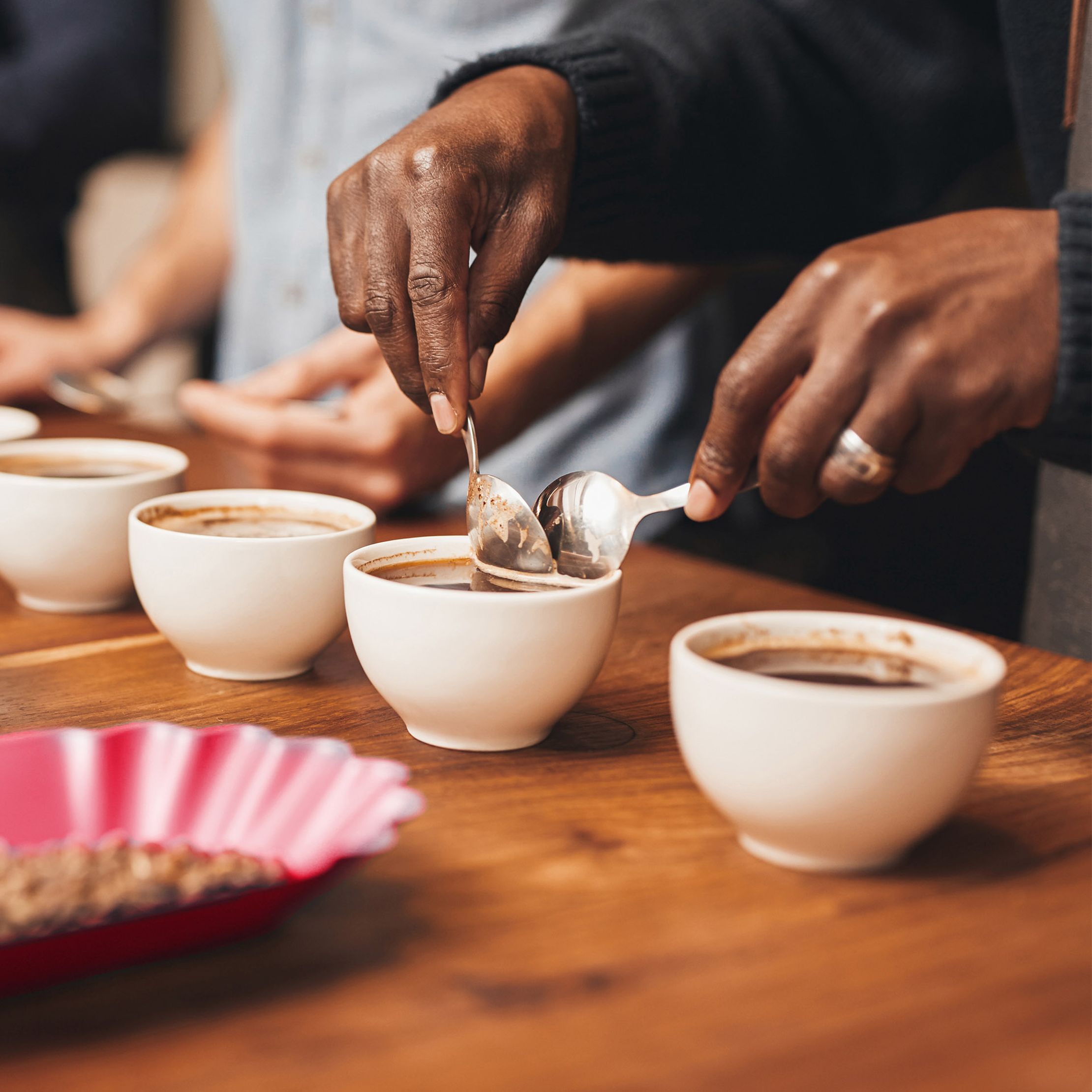 Hands holding spoons over white cups filled with coffee on a wooden table performing cupping