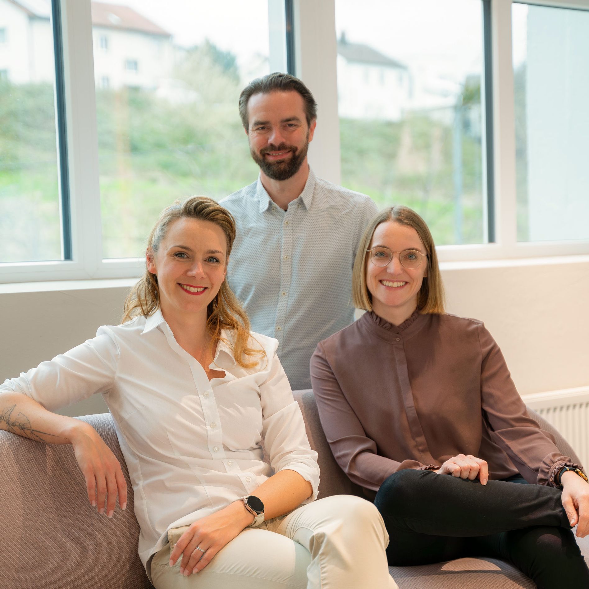 Three people smiling, sitting on a couch in a bright room with large windows and greenery outside.