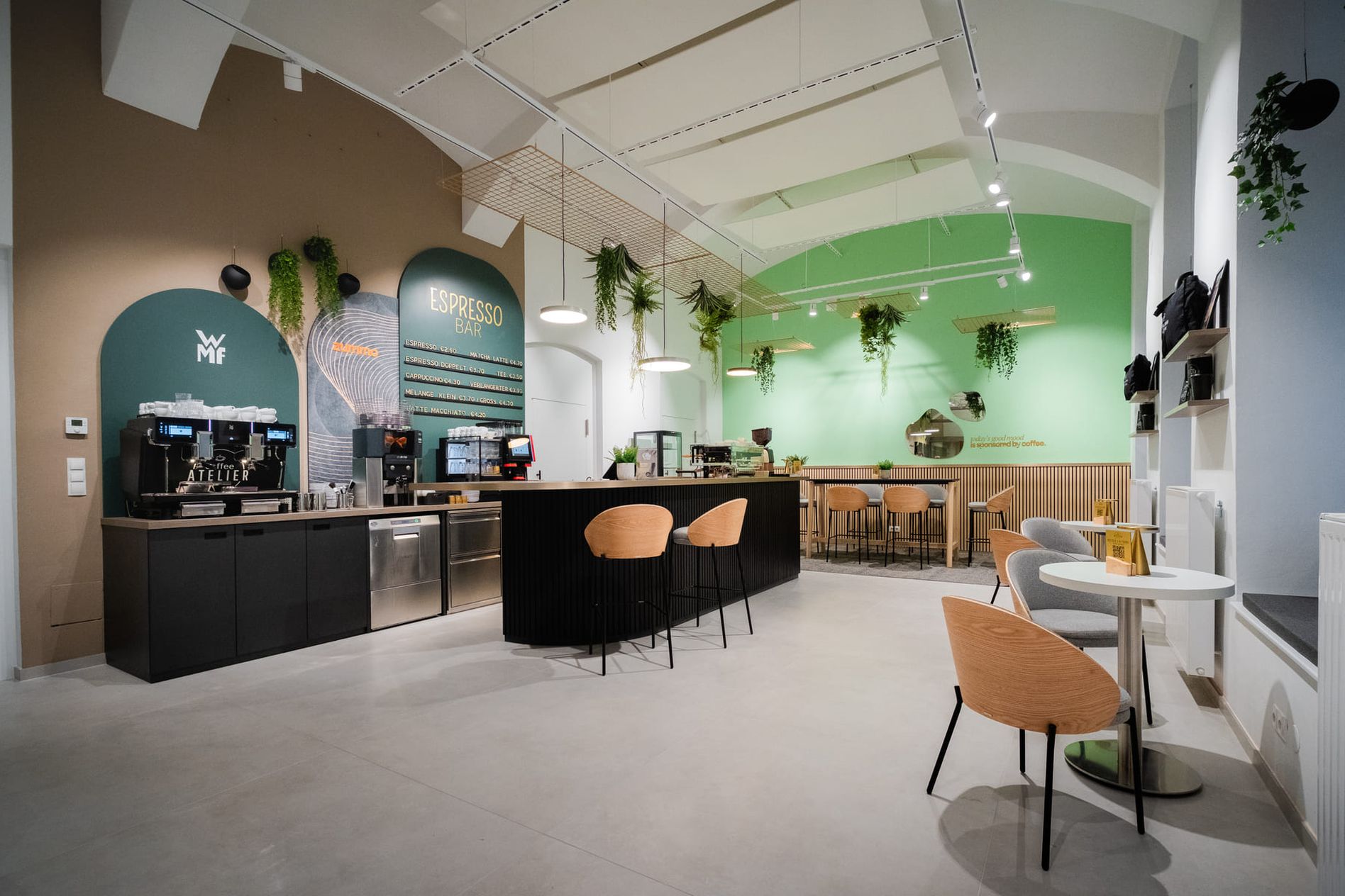 Modern café interior with coffee machines, a wooden counter, green accent walls, hanging plants, and seating areas with wooden chairs and tables.