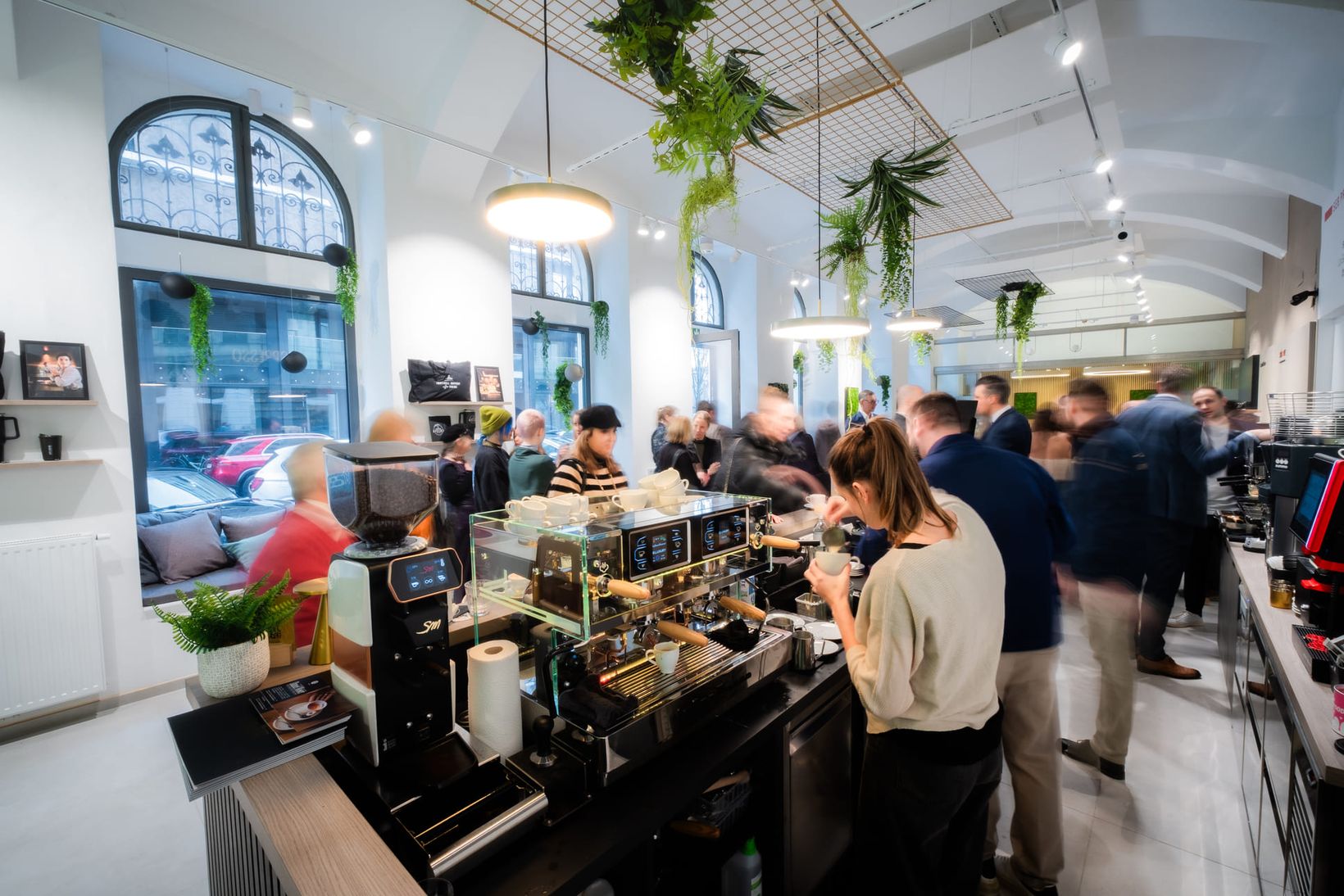 Busy coffee shop with people gathered around professional coffee machines, under modern lighting and greenery, creating a lively atmosphere.