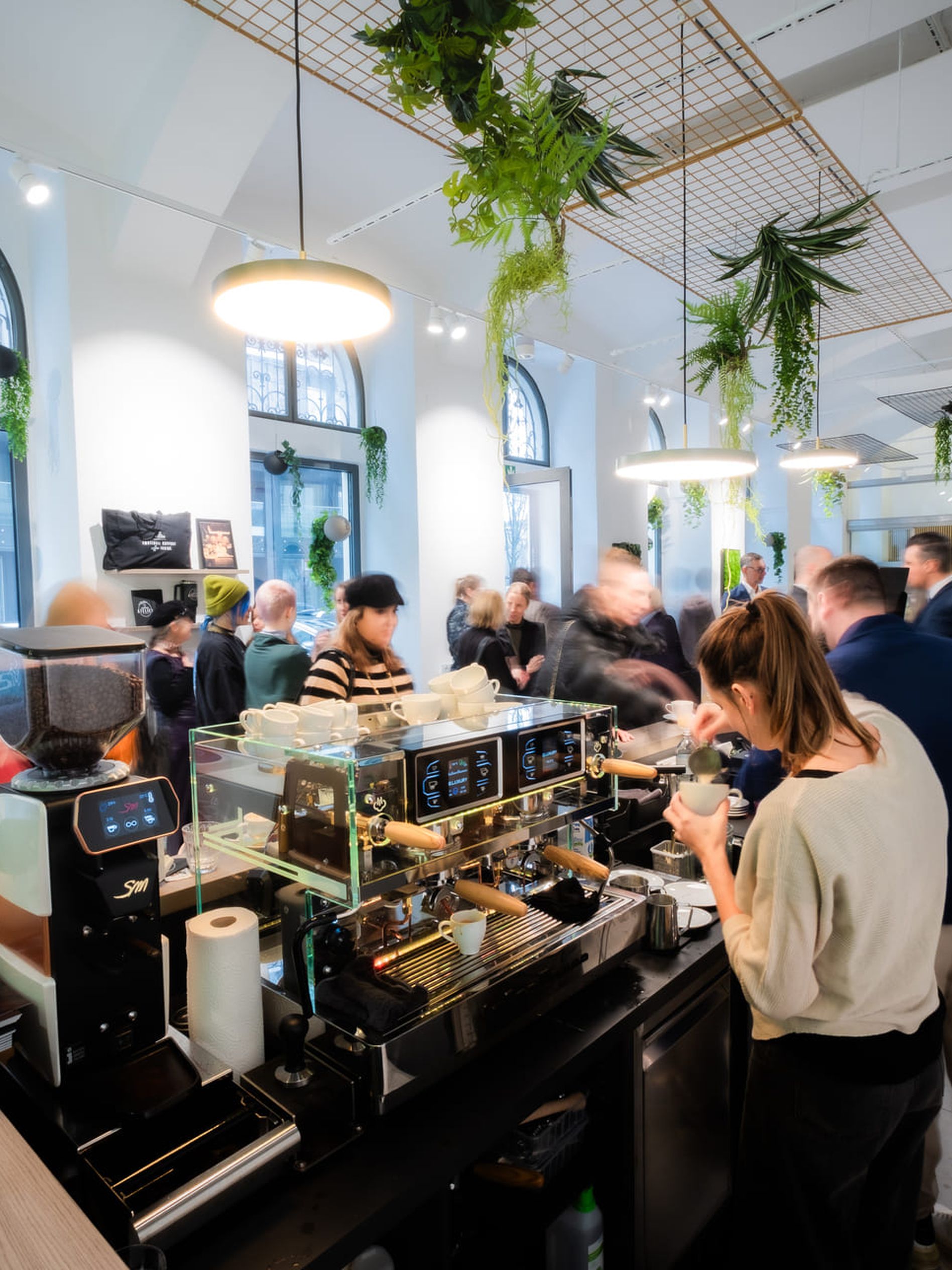 Busy coffee shop with people gathered around professional coffee machines, under modern lighting and greenery, creating a lively atmosphere.