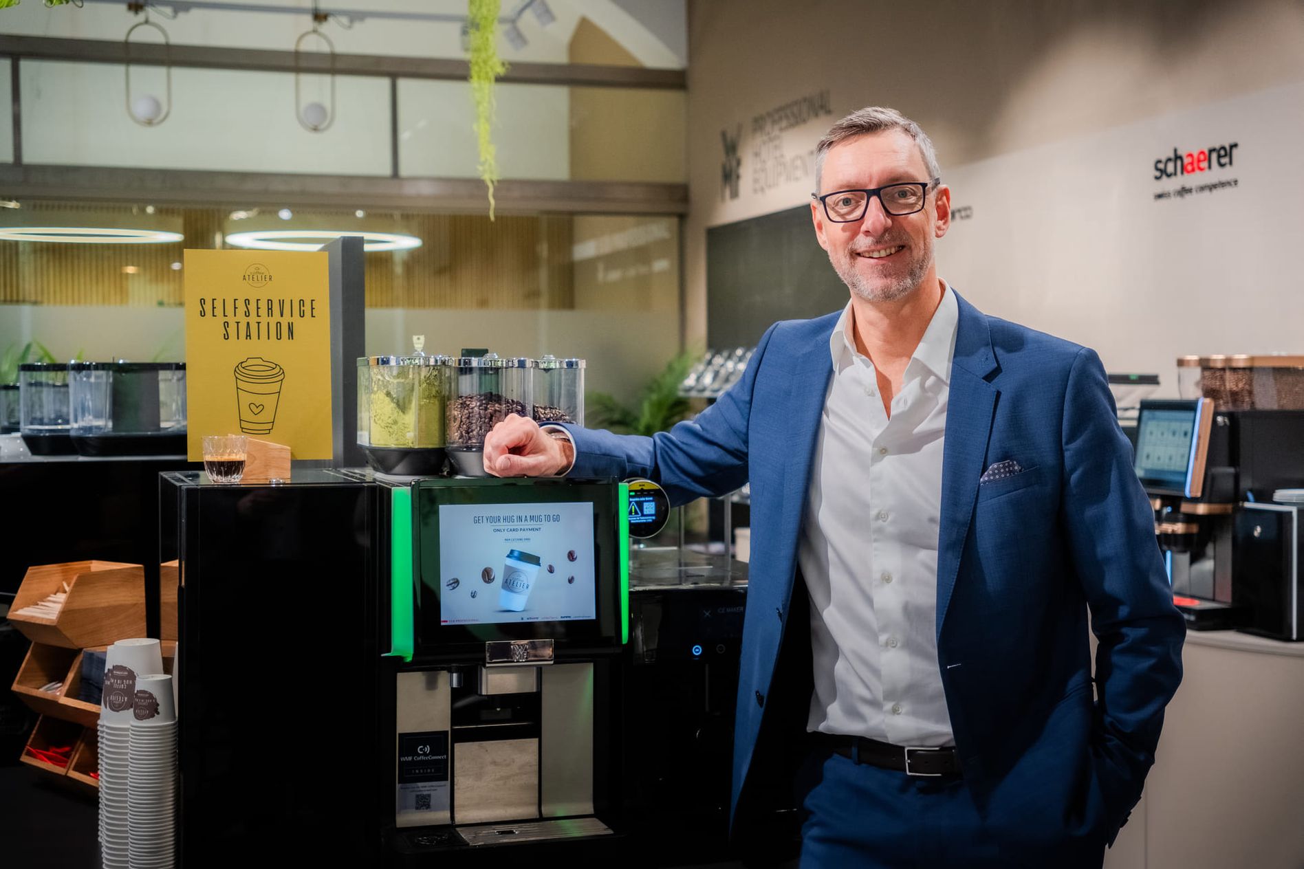 A man in a blue suit stands beside a Schaerer coffee machine at a self-service station in a modern setting.