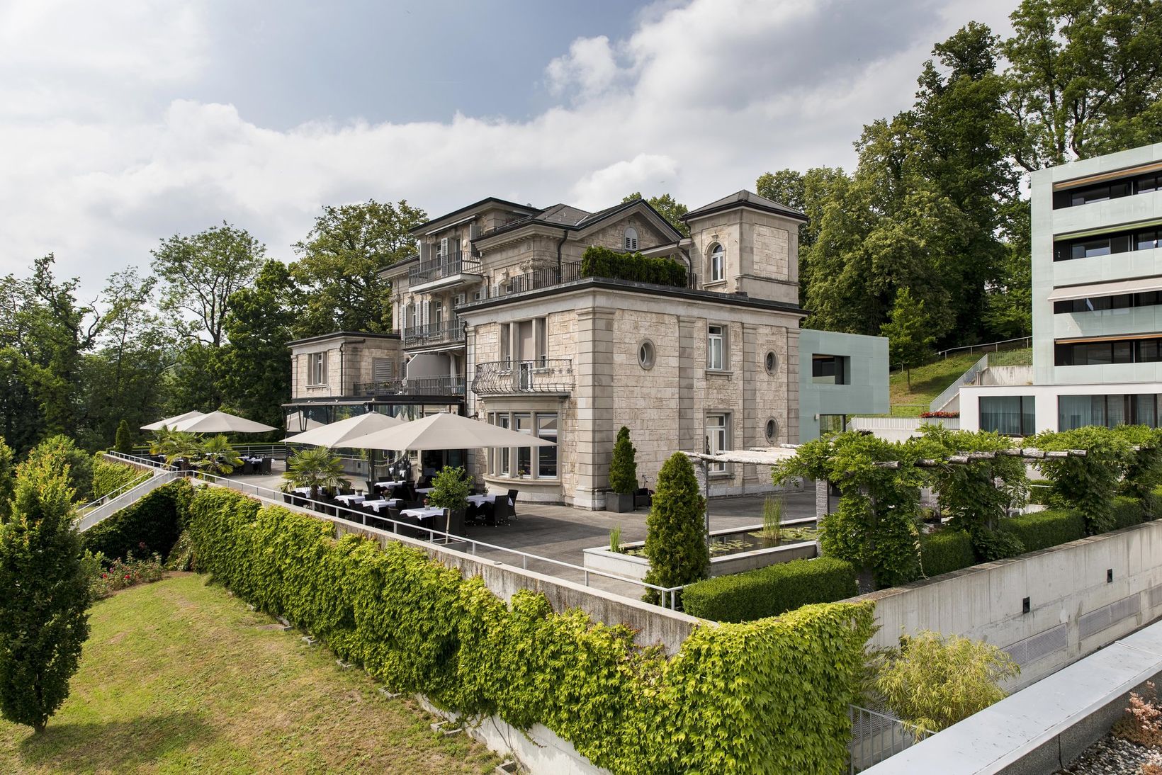 Elegant stone building with terraces, outdoor seating, and greenery, set against a backdrop of trees and a partly cloudy sky.
