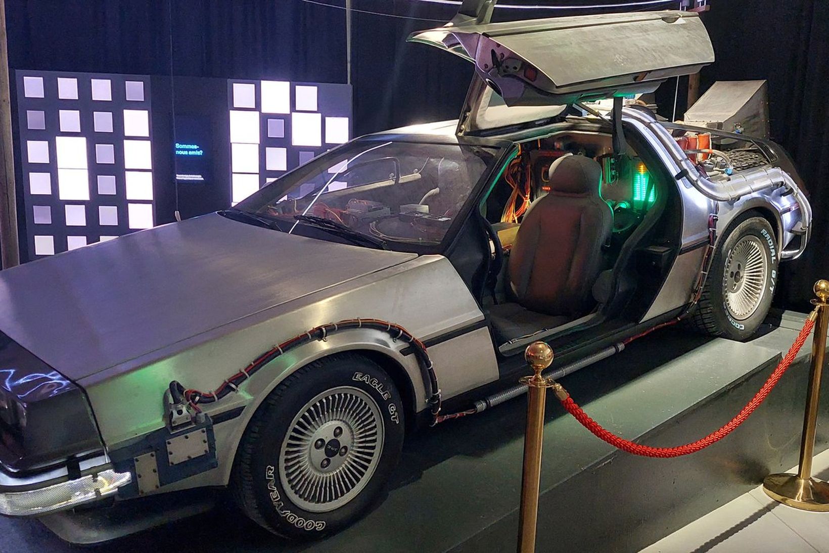 A silver DeLorean car with gull-wing doors open, displayed on a platform with red ropes, in a dimly lit exhibition space.