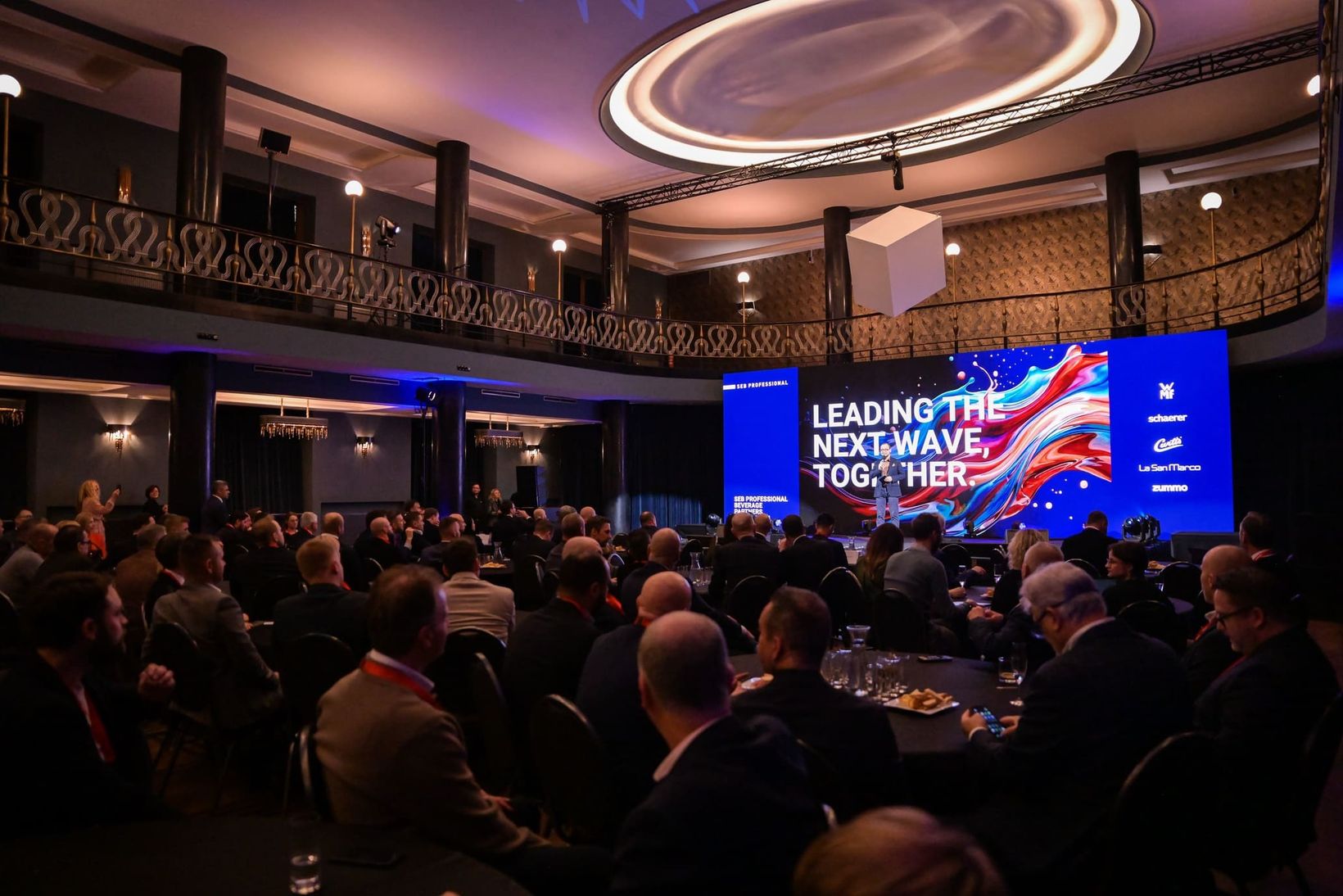Conference with audience seated, facing a large screen displaying "LEADING THE NEXT WAVE, TOGETHER" in an elegant, modern venue.