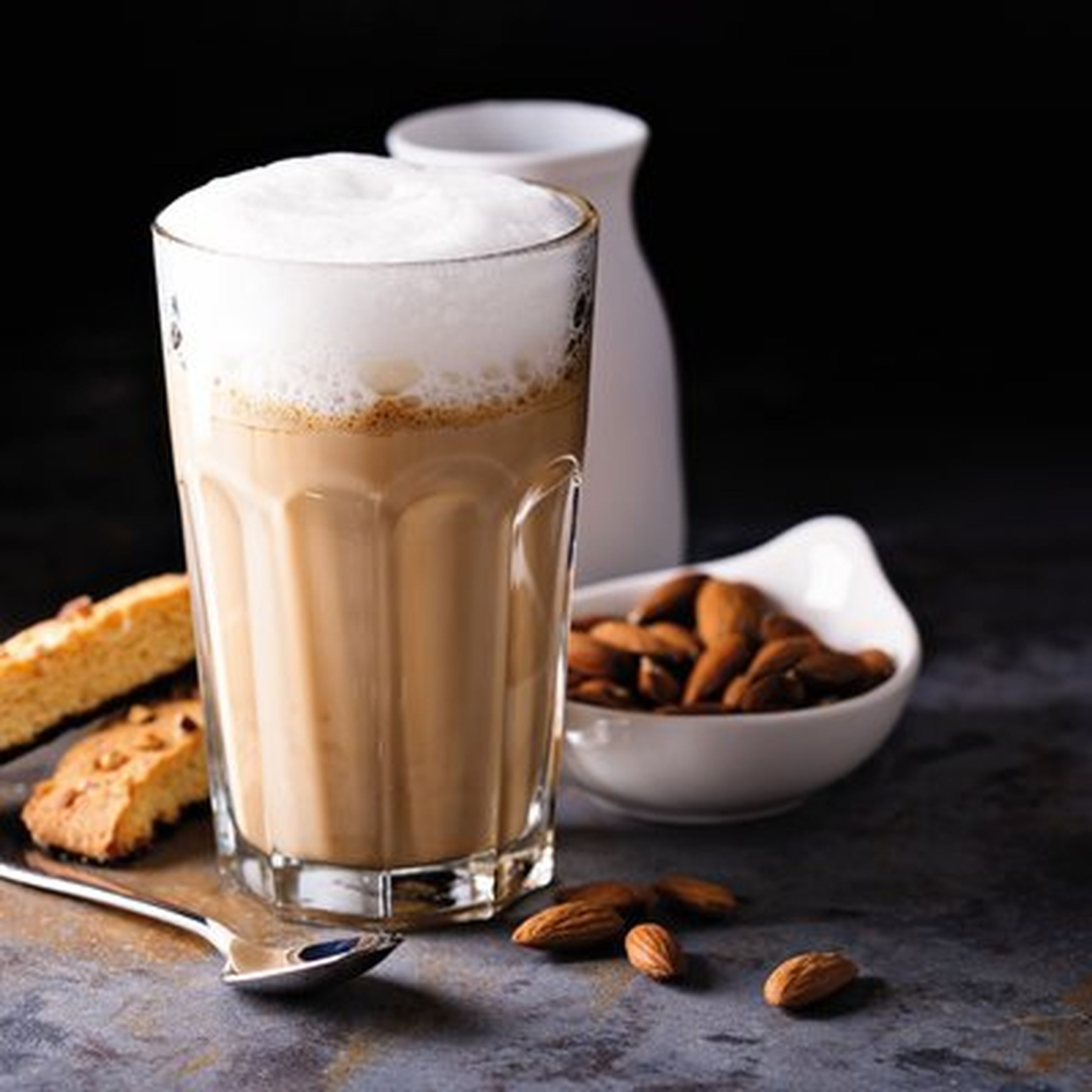 A frothy latte in a glass, accompanied by biscotti, almonds in a bowl, and a small white pitcher on a dark surface.