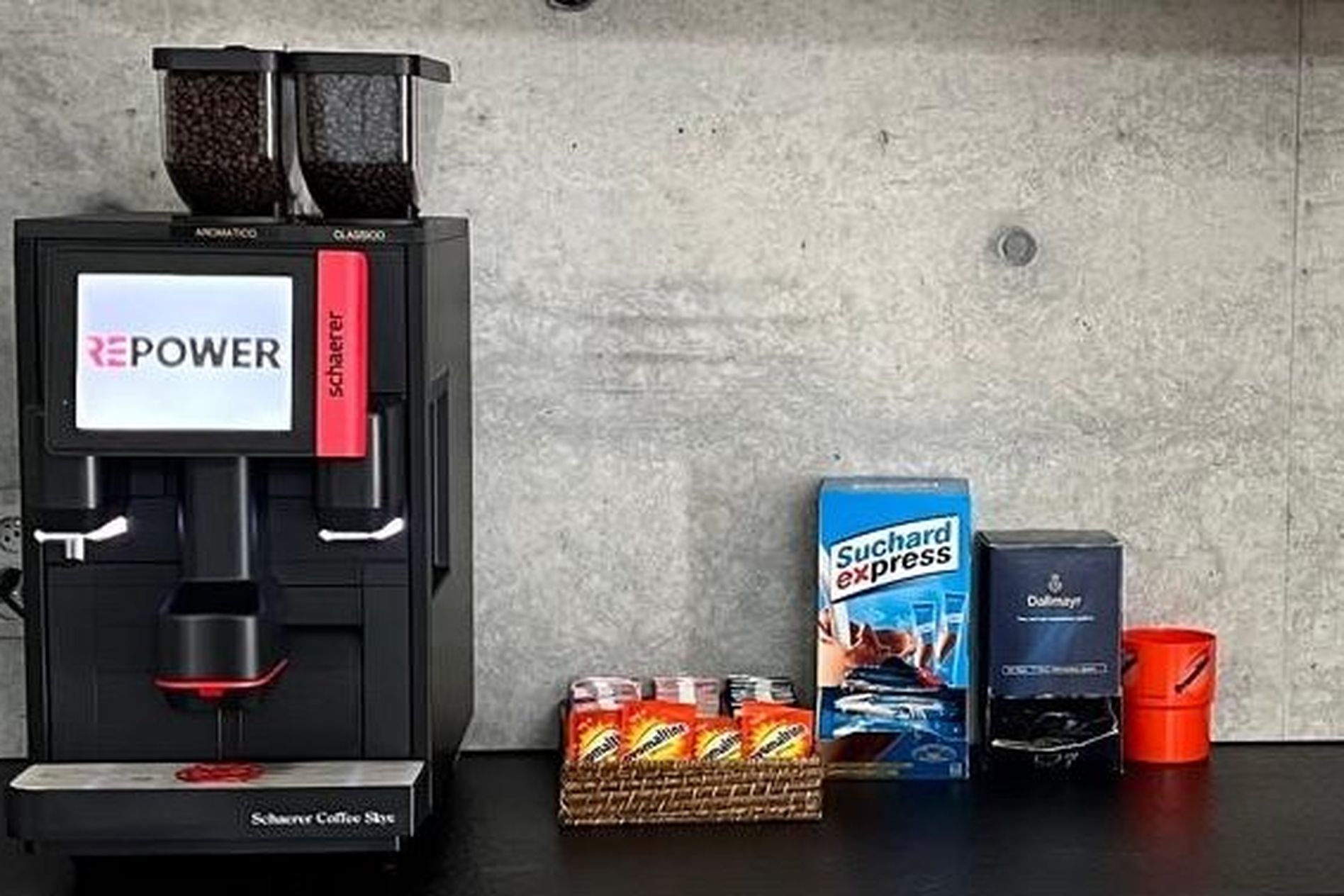 Coffee machine on a counter with various drink supplies, including instant coffee packets, sugar, and a red mug against a gray wall.