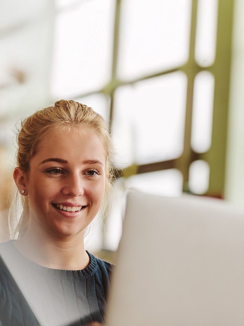 Smiling woman with blonde hair working on a laptop in a bright room with large windows.