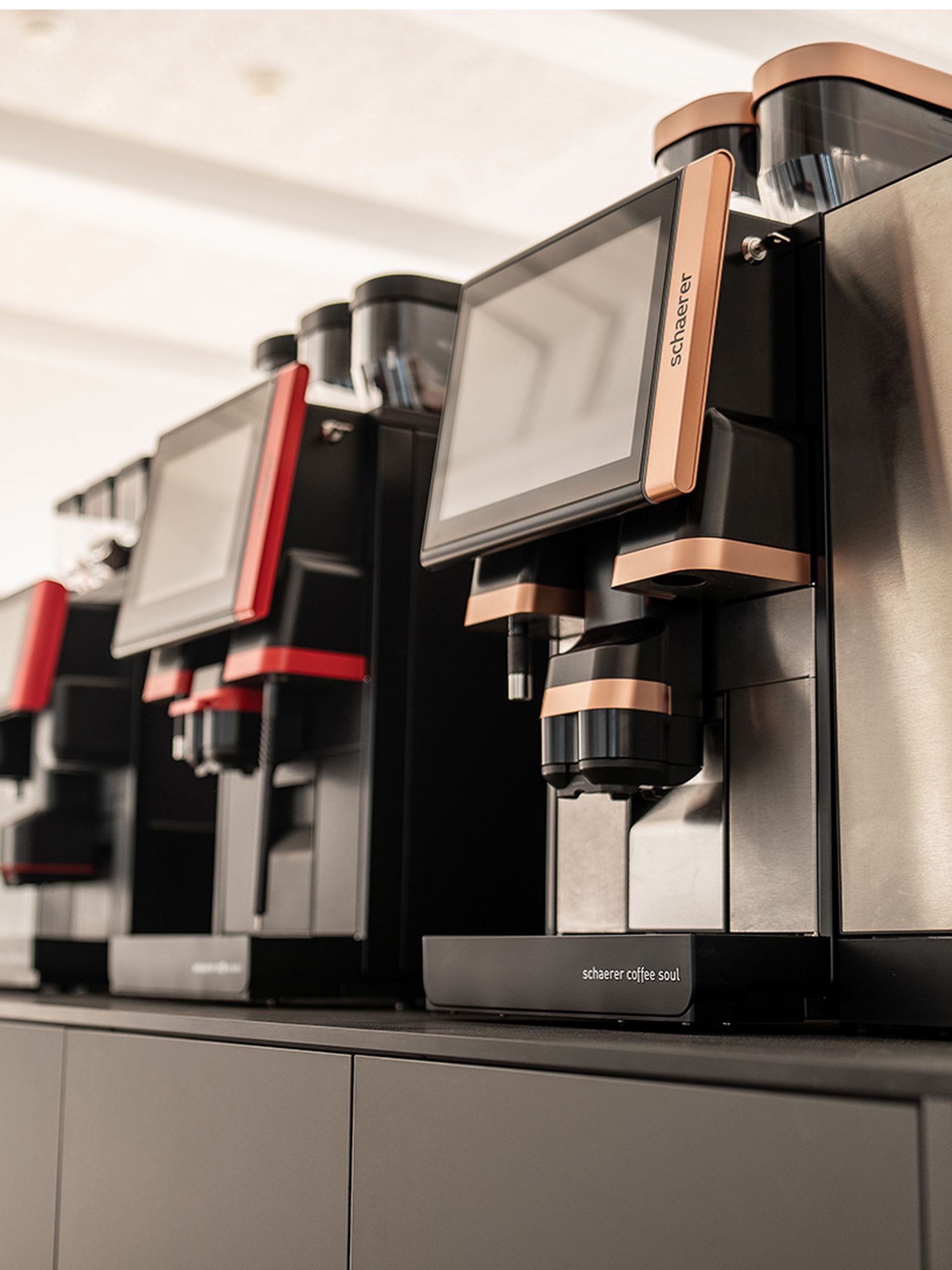 Close-up of sleek, modern coffee machines with digital screens and copper accents, lined up on a counter in a well-lit space.