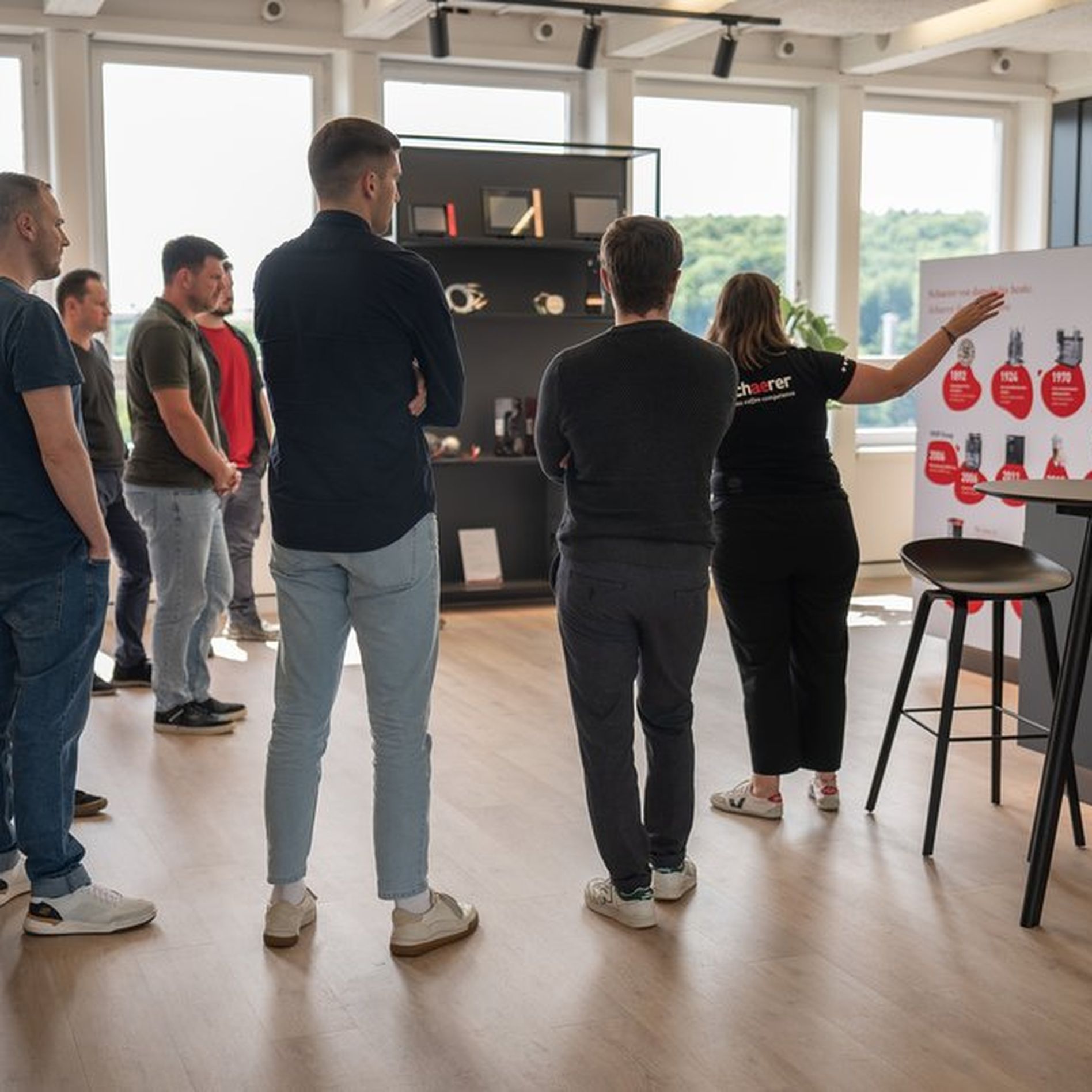 A group of people stands in an office, listening to a presentation near a display board with diagrams and images. Large windows are in the background.