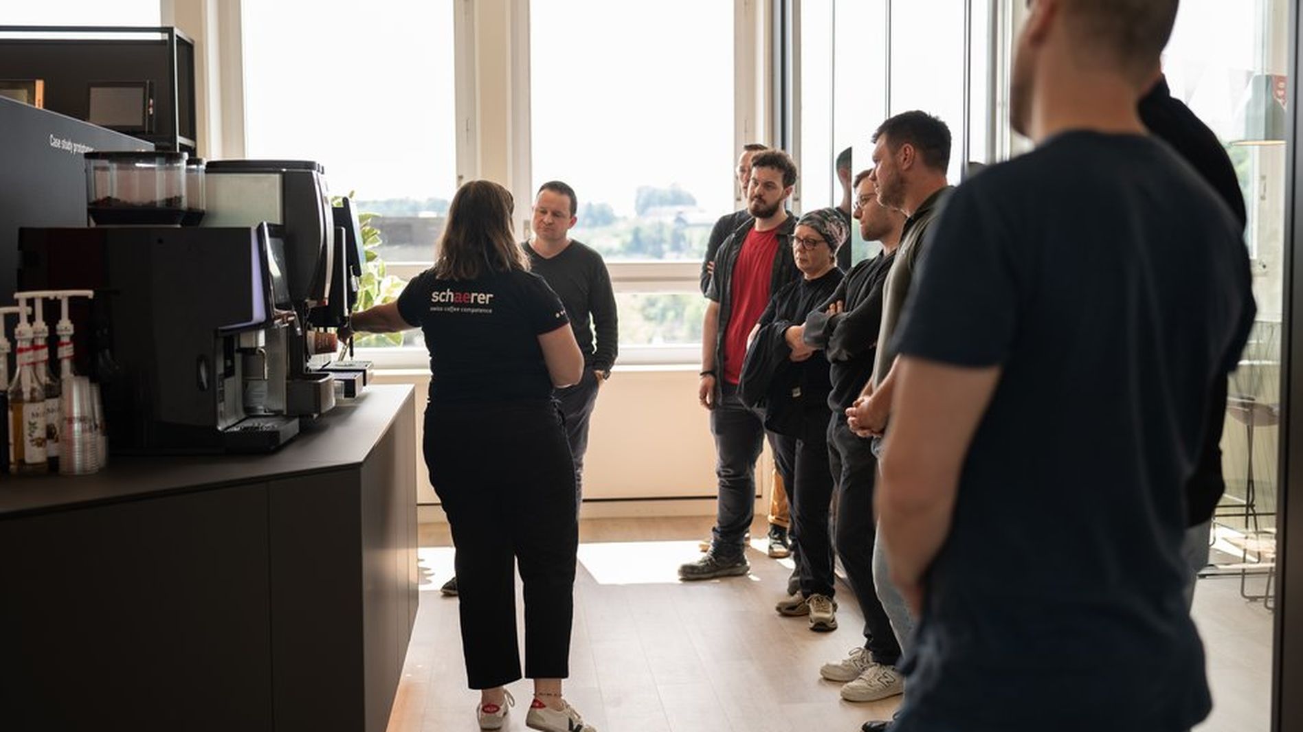 A group of people stands attentively in an office as a woman demonstrates a coffee machine. Large windows provide a bright, open atmosphere.