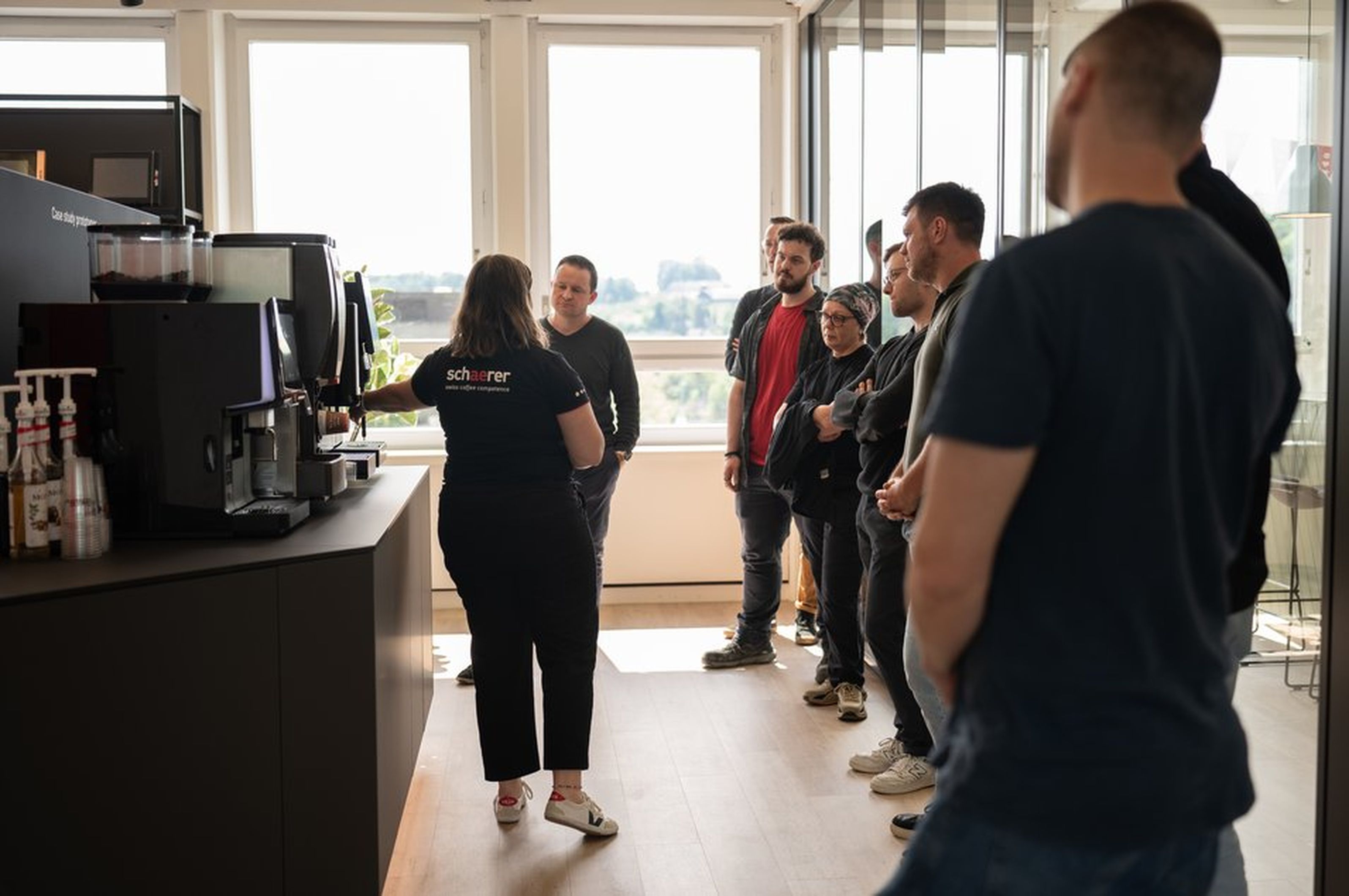 A group of people stands attentively in an office as a woman demonstrates a coffee machine. Large windows provide a bright, open atmosphere.
