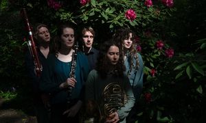 Five musicians stand with woodwind instruments among lush greenery and pink flowers, softly illuminated by dappled sunlight.