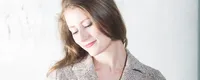 Woman in a tweed jacket gently holding a violin, looking down with a serene expression against a light background.