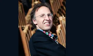 Smiling person wearing glasses and a colorful shirt, standing in front of an organ.
