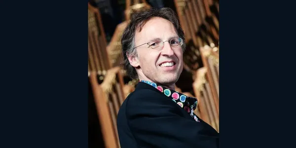 Smiling person wearing glasses and a colorful shirt, standing in front of an organ.